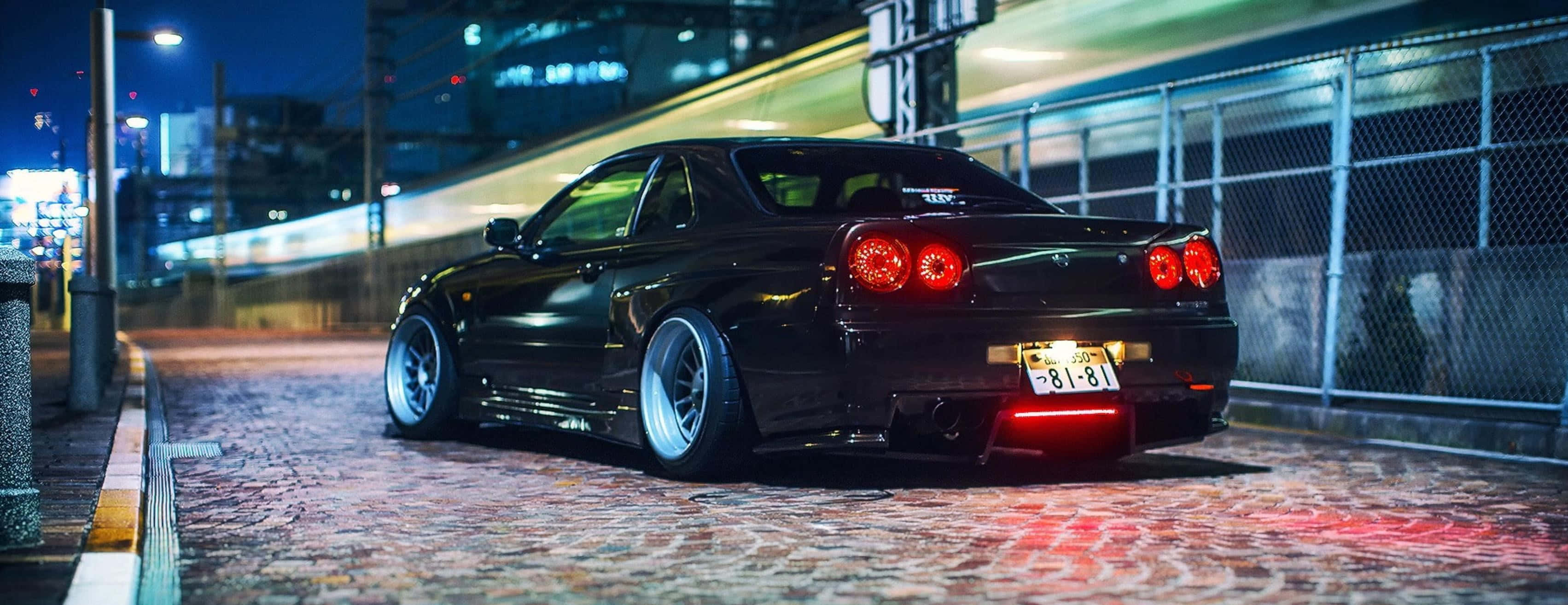 傳奇R33 GTR 壁紙 — 一輛時尚的黑色跑車，帶有發光的紅色尾燈，停在城市夜晚的鵝卵石街道上。