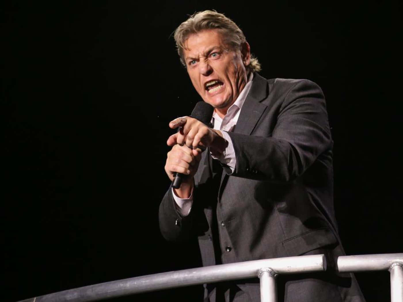 傳奇WWE摔角手威廉·雷加爾於WWE活動中激昂演講 壁紙 — 一名身穿西裝的男子在黑暗的舞台上，在強烈的燈光下熱情地對著麥克風唱歌。