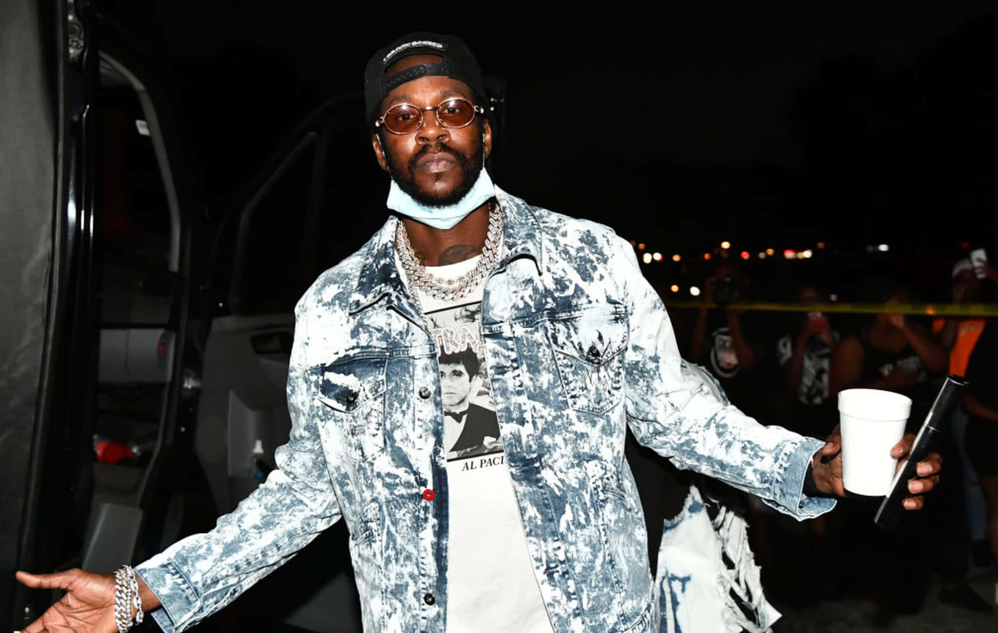 傳奇嘻哈藝人2 Chainz 壁紙 — 一個穿著破舊牛仔外套和帽子的男人在夜晚自信地站著，手拿杯子和電話，背後的城市燈光閃爍。