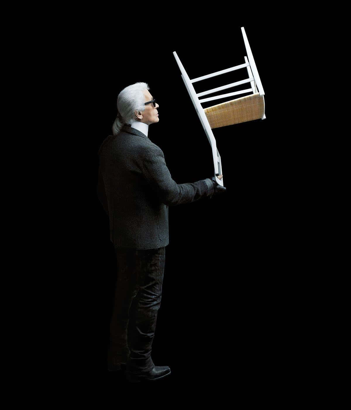 傳奇時裝設計師Karl Lagerfeld為鏡頭擺姿 壁紙 — 一名身穿深色西裝、戴著太陽眼鏡的男子，在黑色背景前倒舉著一張白色椅子。