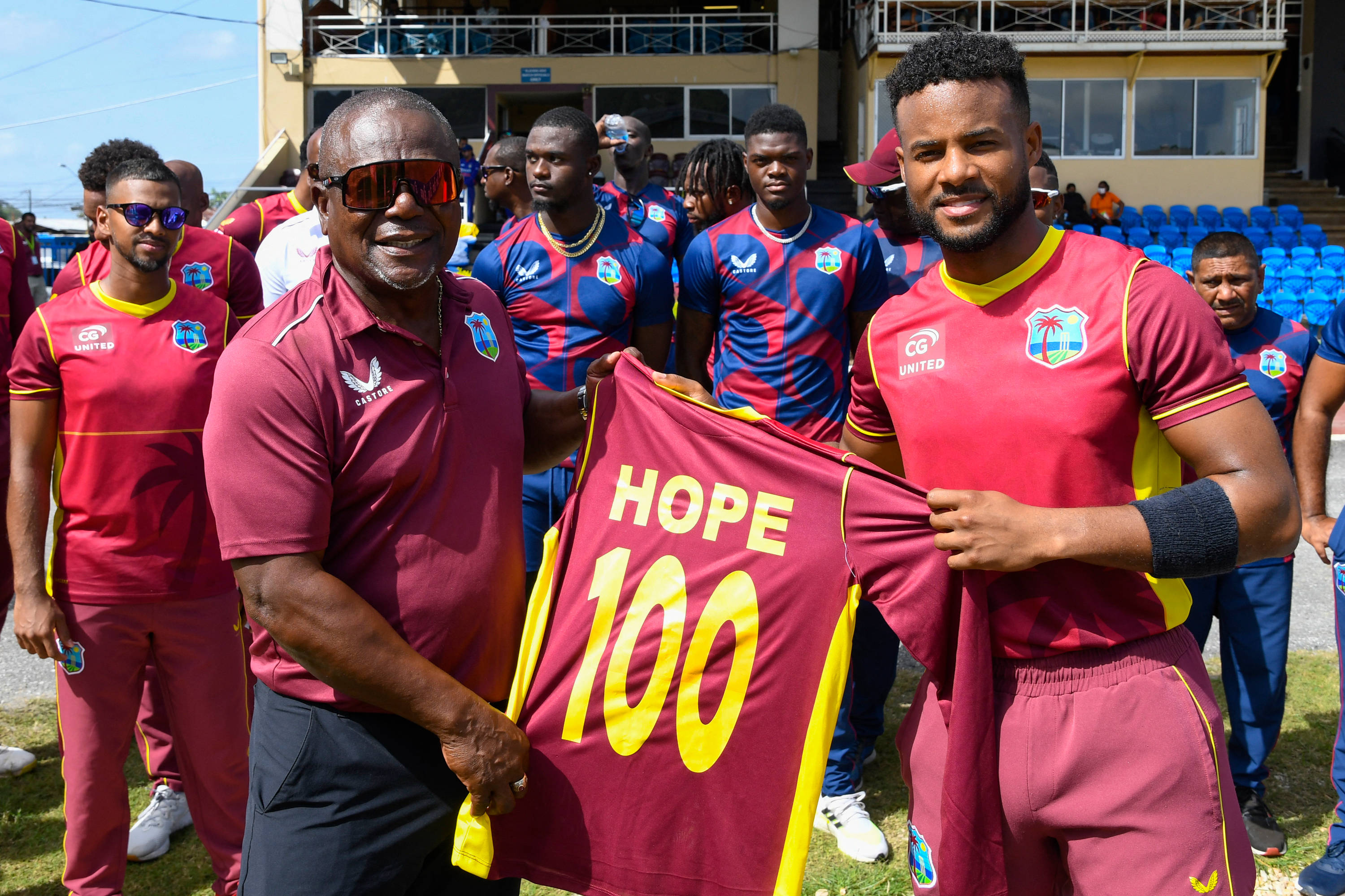 傳奇板球偶像 Shai Hope 與 Desmond Haynes 壁紙 — 兩名微笑的男子身穿栗色運動服，高舉一件印有HOPE 100字樣的球衣，隊友們站在他們身後，背景是陽光明媚的球場。