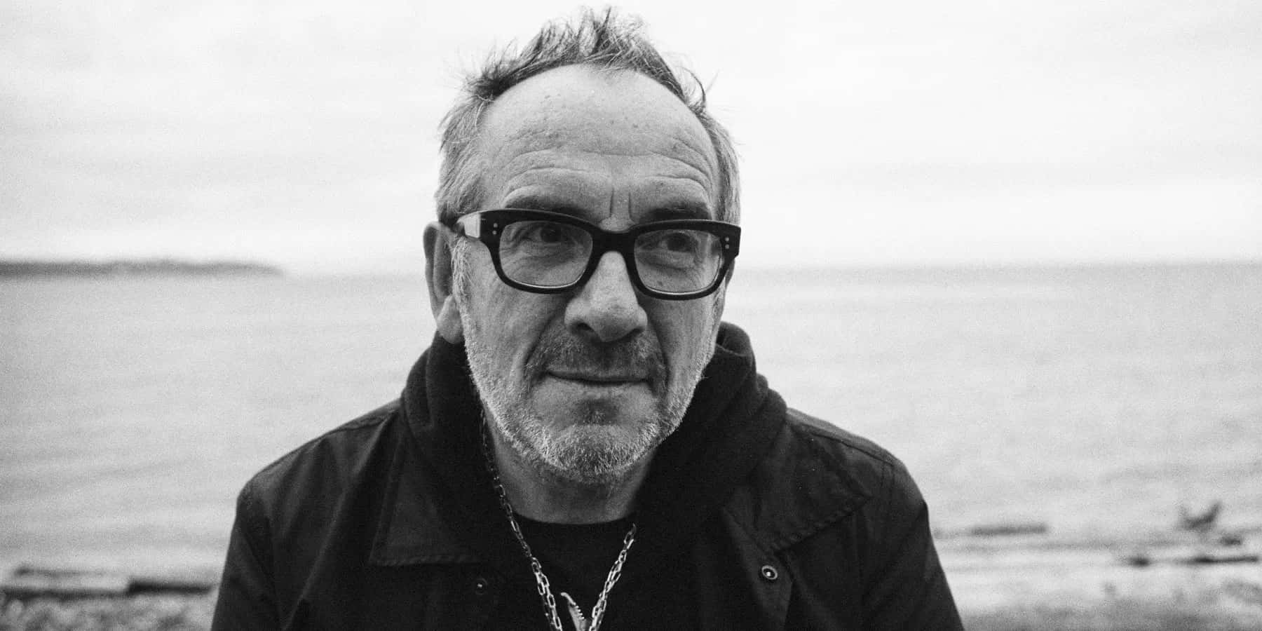 傳奇歌手Elvis Costello 壁紙 — 一個戴眼鏡、留鬍鬚的男人站在水邊的黑白肖像。