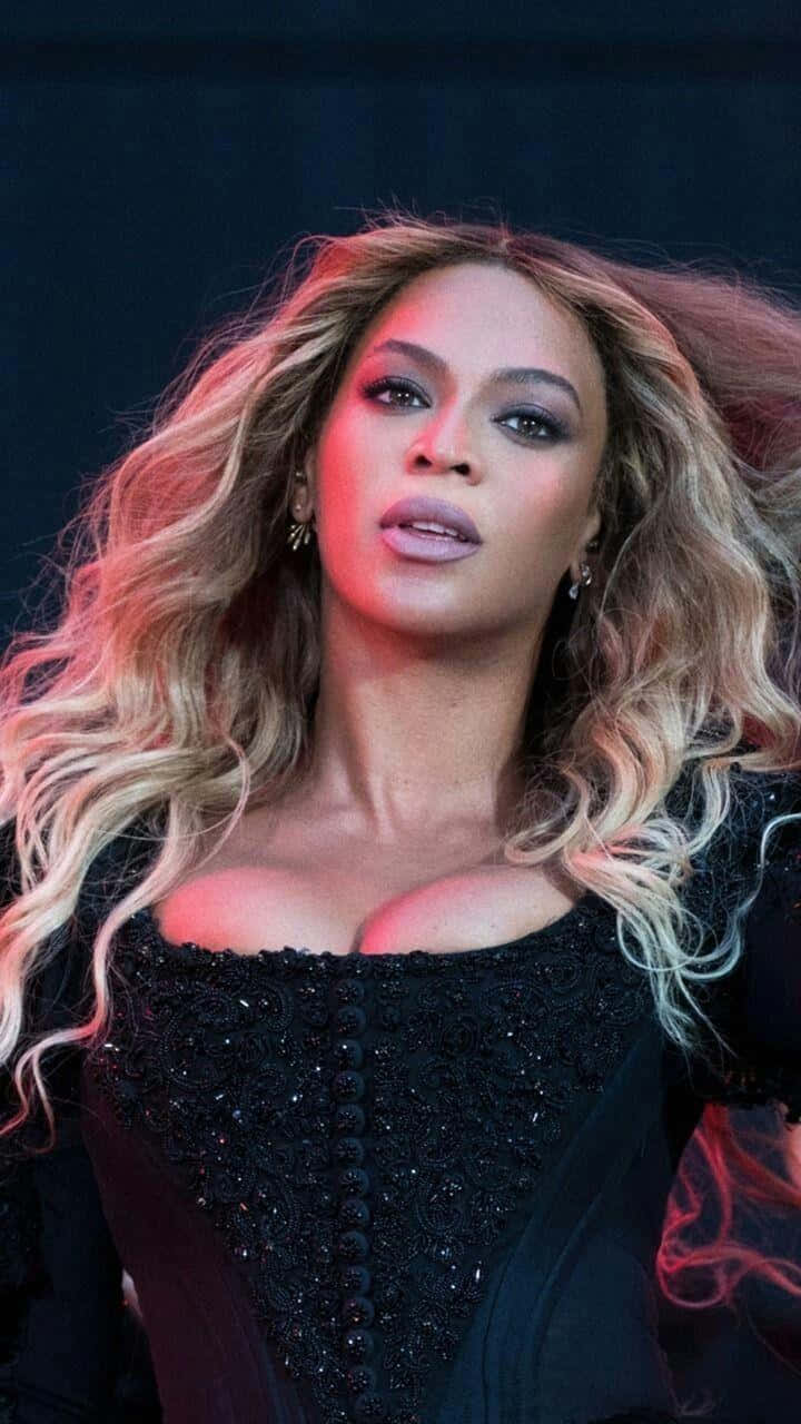 傳奇歌手碧昂絲 壁紙 — Beyonce 一頭飄逸金髮，在紅色燈光下，身穿黑色鑲飾上衣，在深色背景中凝視。