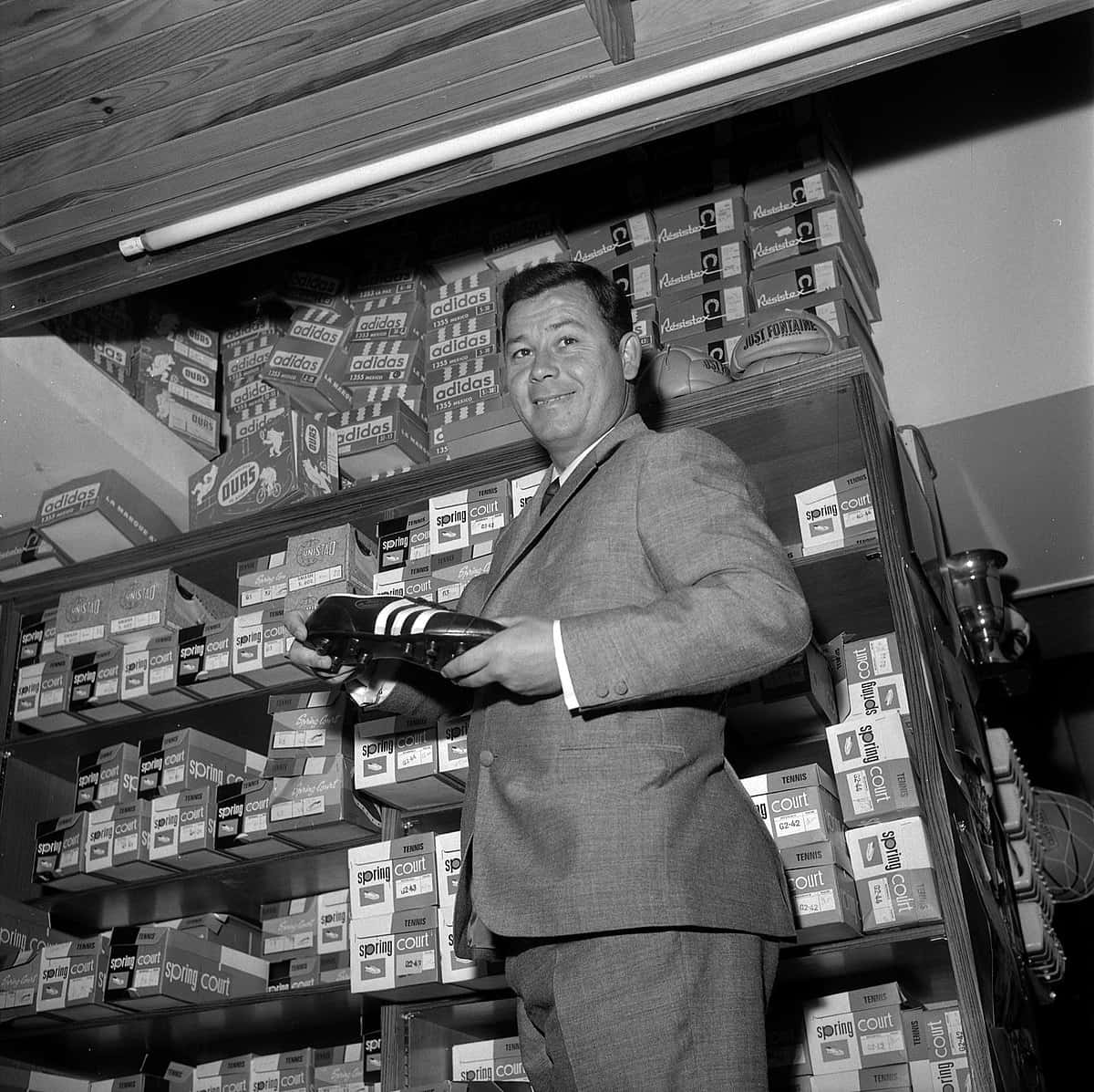 傳奇法國足球員Just Fontaine 壁紙 — 一位微笑的西裝男子在充滿品牌盒子的復古鞋店中拿著一雙黑色Adidas鞋。