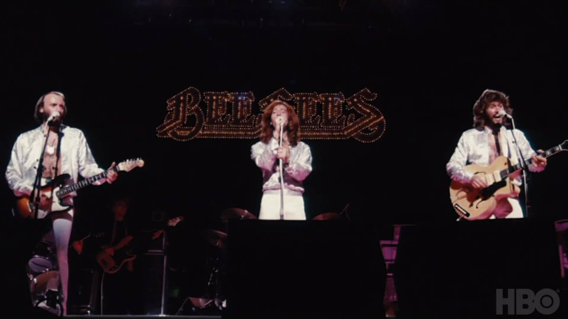 傳奇流行樂團 Bee Gees 演唱會現場 壁紙 — 三位身穿白色服裝的音樂家在閃閃發光的 Bee Gees 標誌下，手持結他和咪高峰在舞台上表演。