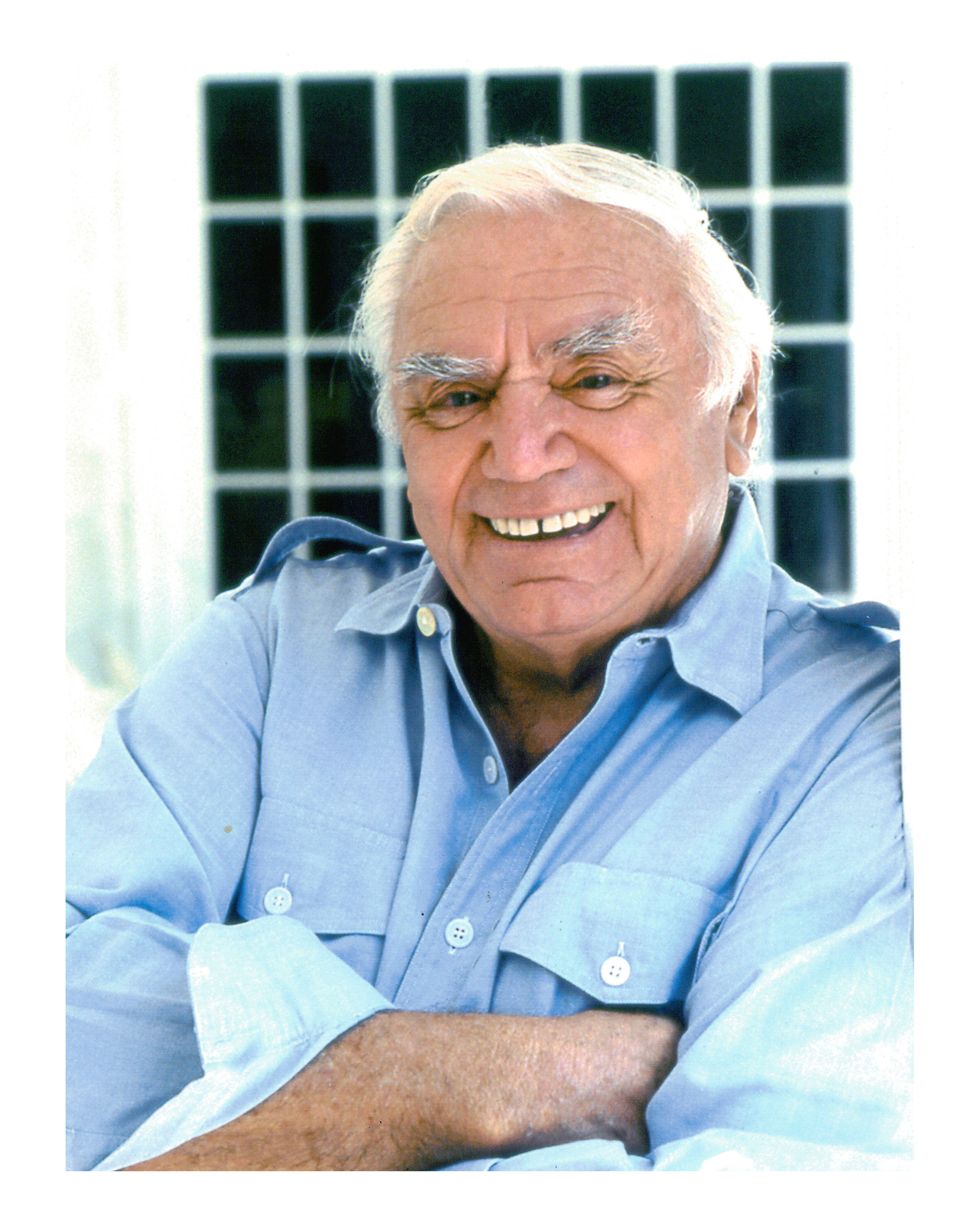 傳奇演員 Ernest Borgnine 穿藍襯衫 壁紙 — 一位白髮老人穿著淺藍色襯衫，在深色格窗背景下溫暖地微笑。