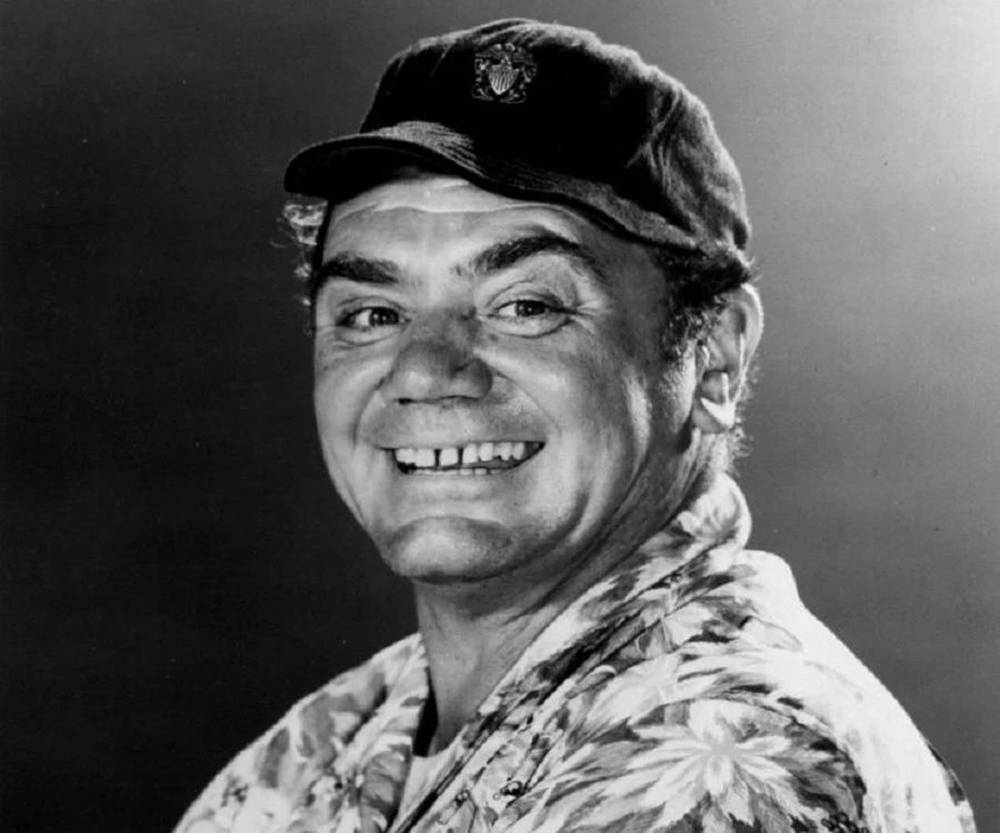傳奇演員 Ernest Borgnine 戴棒球帽 壁紙 — 一個戴帽同花恤衫嘅開心男人喺黑色攝影棚背景前露出燦爛笑容。