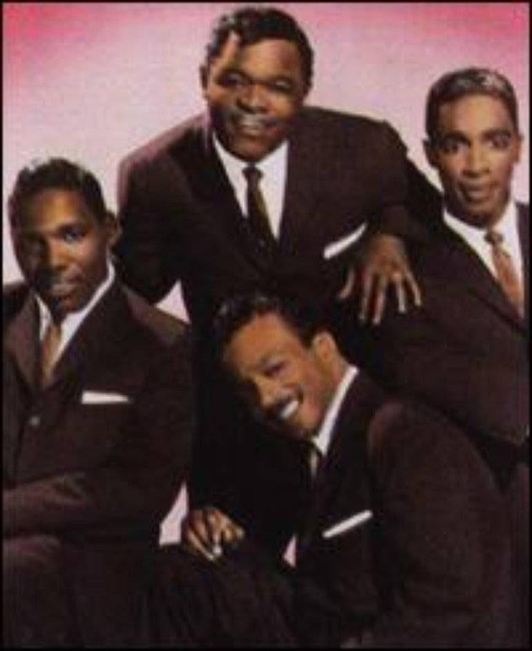 傳奇美國樂團 The Drifters 演唱會現場 壁紙 — 四位微笑的黑人男士穿著俐落西裝，在柔和的粉紅色漸變背景前動感地擺姿勢。