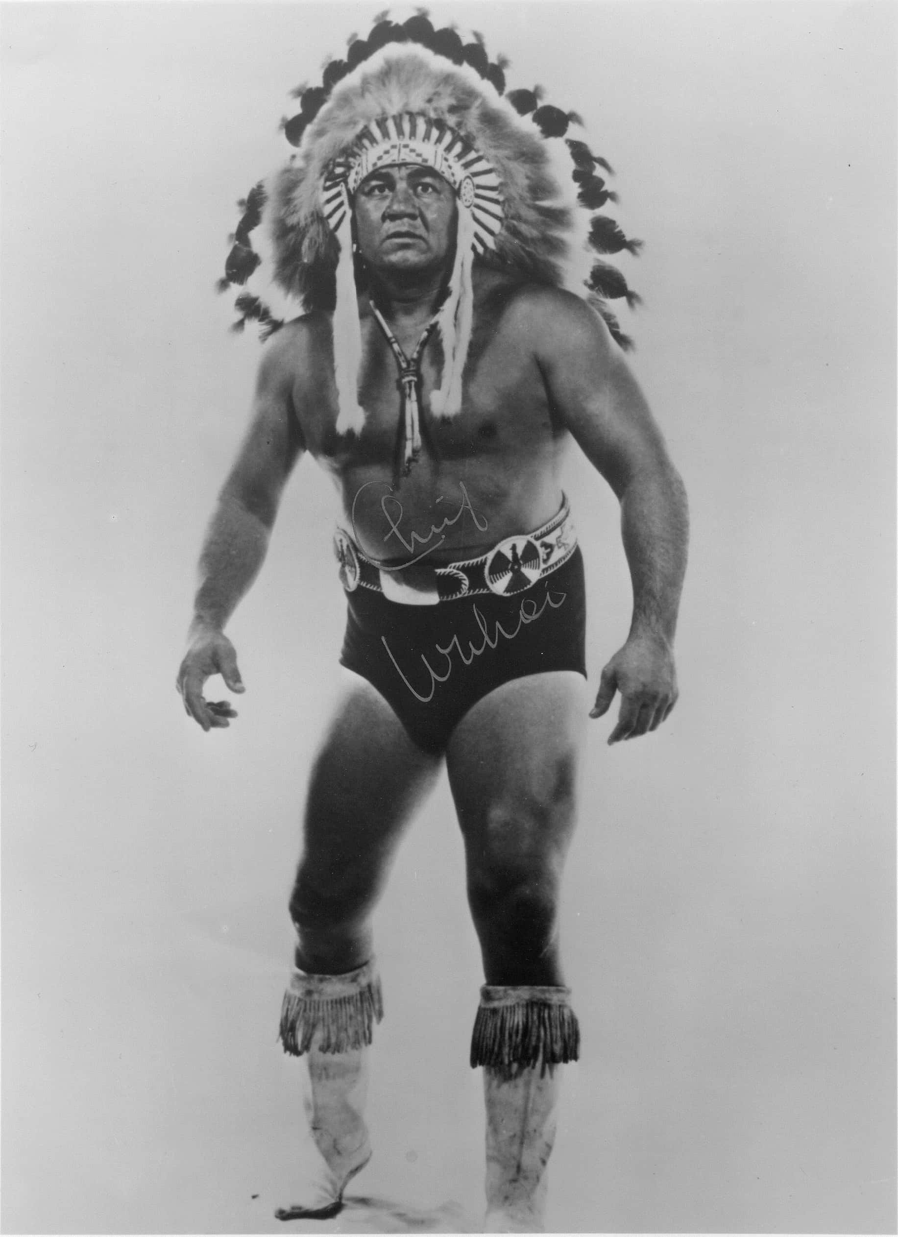 傳奇美洲原住民摔角手Wahoo McDaniel經典影像 壁紙 — 一位肌肉發達的男子，戴著羽毛頭飾和流蘇短褲，在工作室肖像中自信地站立。