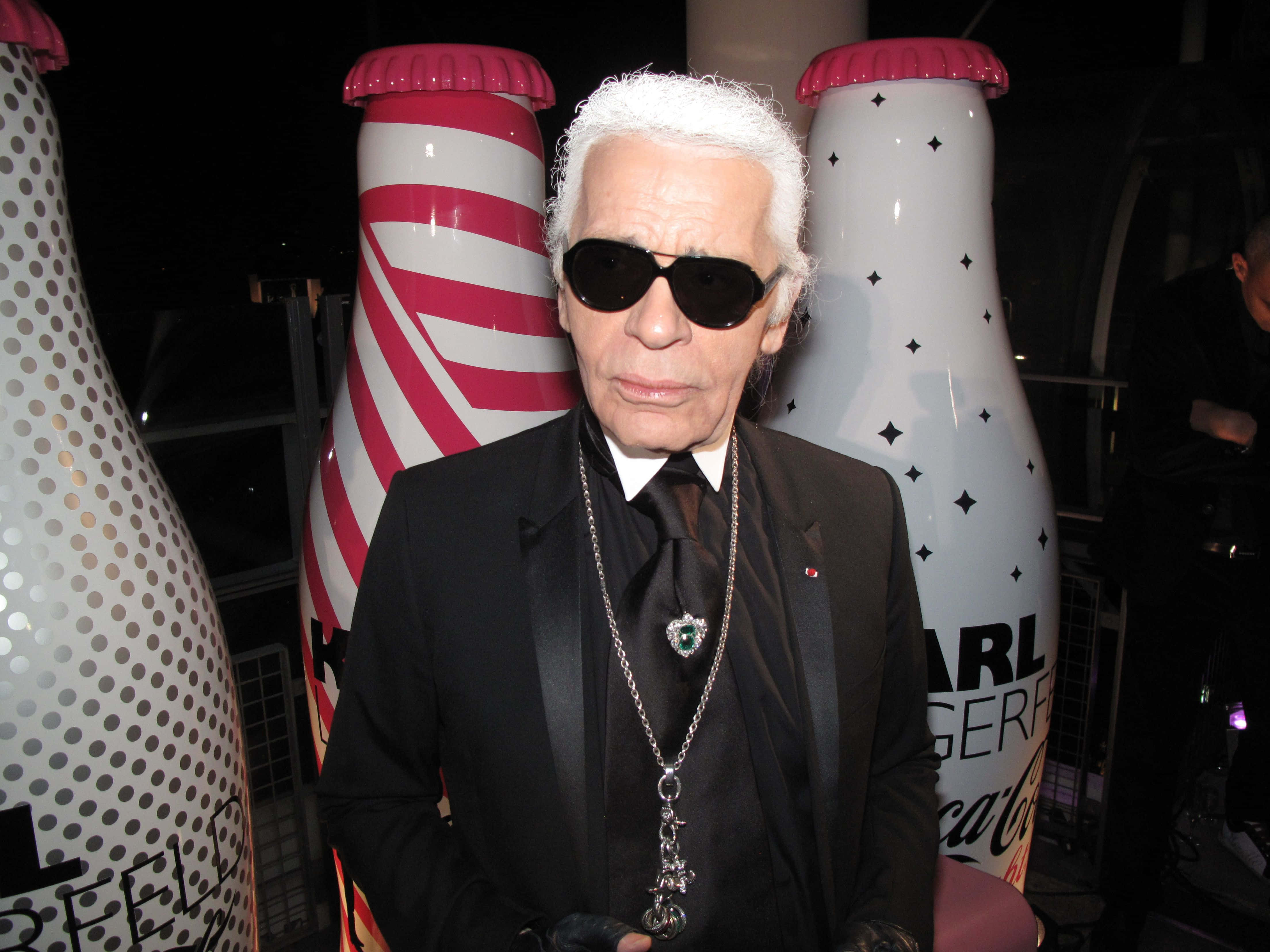 傳奇設計師 Karl Lagerfeld 壁紙 — 卡爾·拉格斐身穿黑色西裝和太陽眼鏡，在夜間活動中站在巨大的風格化可口可樂瓶前。