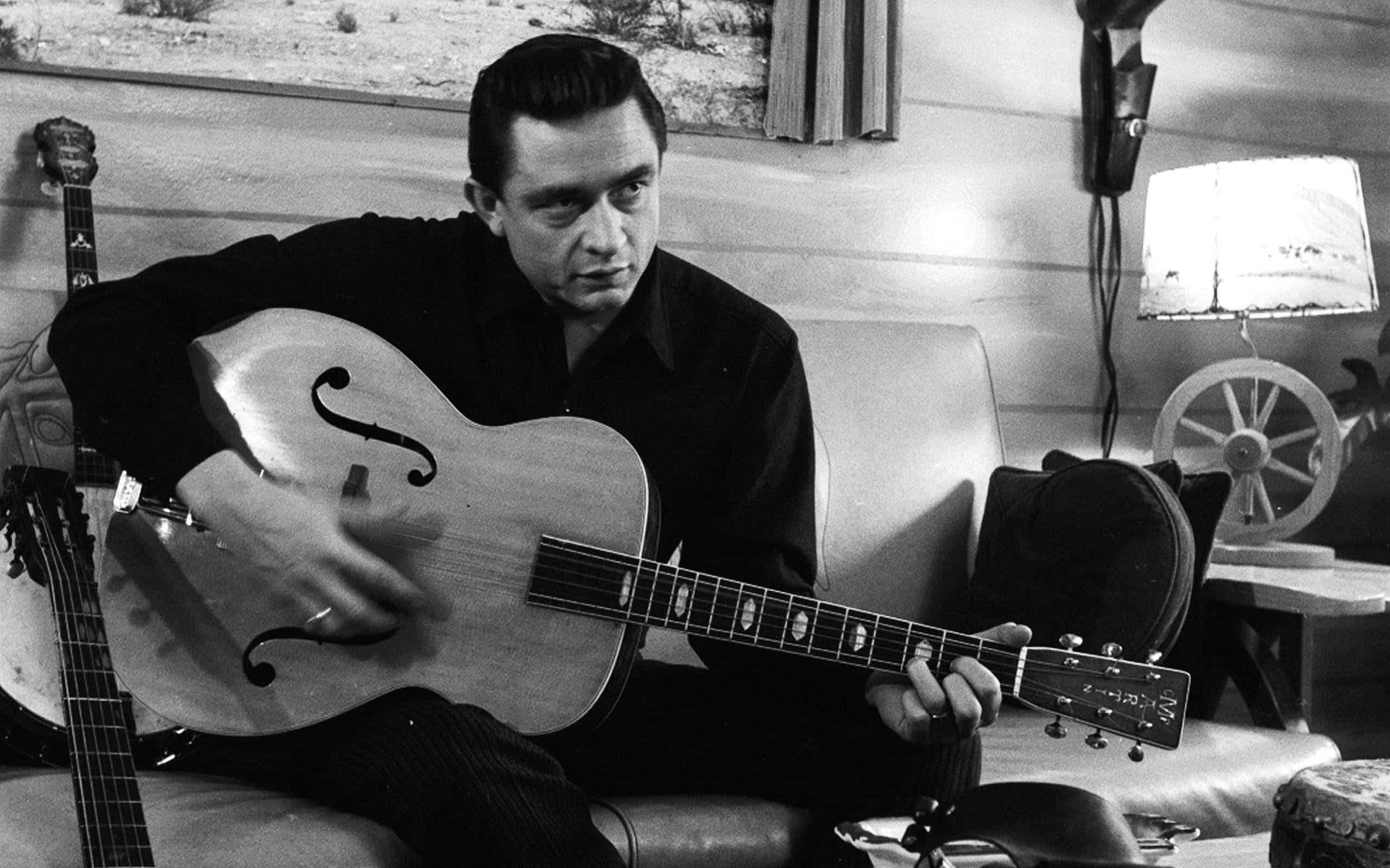 傳奇音樂人 Johnny Cash 壁紙 — 一個男人坐在沙發上，拿著一把空心結他，在一個溫暖燈光的復古房間裡，有木牆和一盞燈。