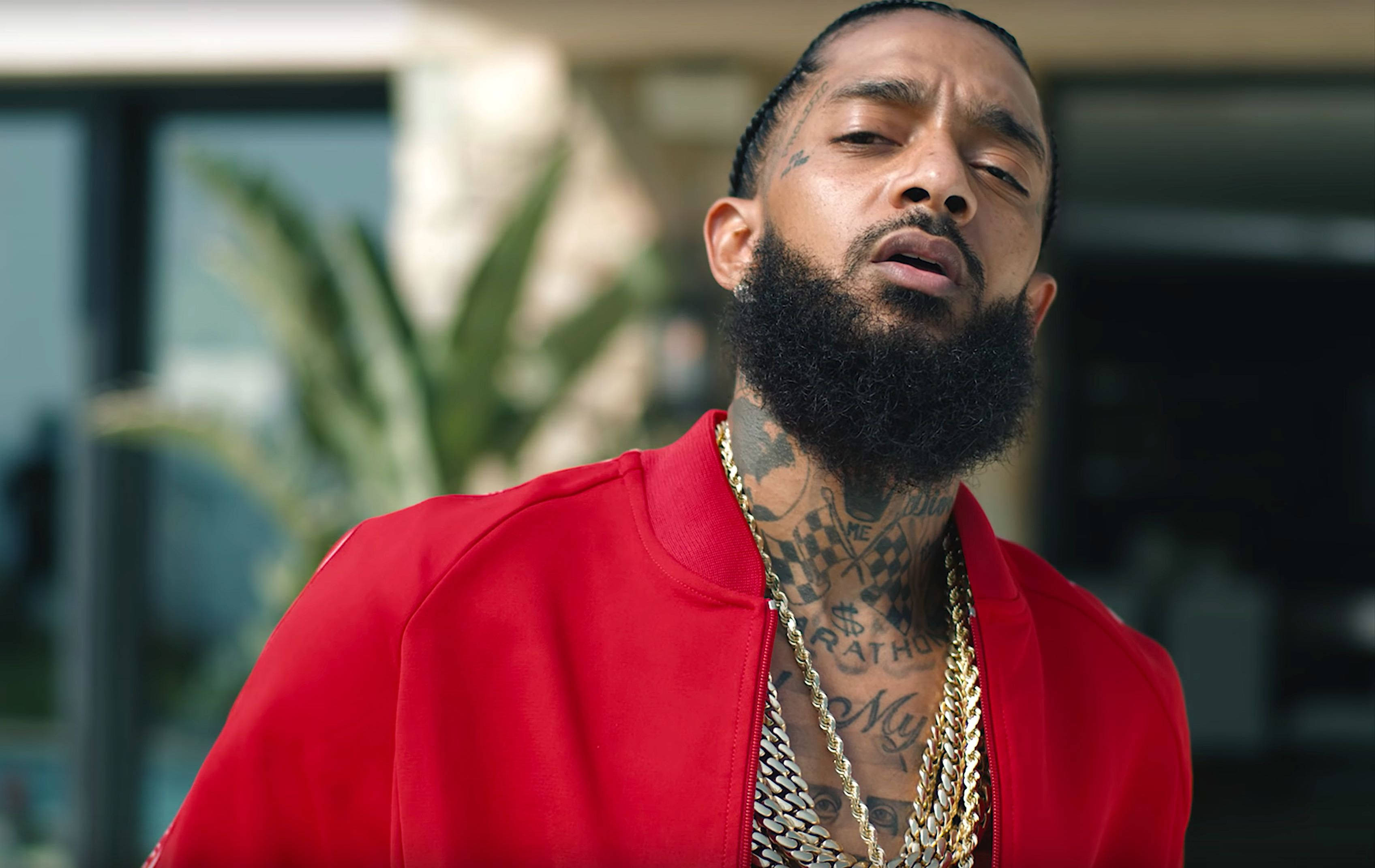 傳奇饒舌歌手 Nipsey Hussle 在音樂工作室散發獨特魅力 壁紙 — 一位留鬍鬚及有紋身的男士穿著紅色外套和戴著金鏈，在戶外向上望。