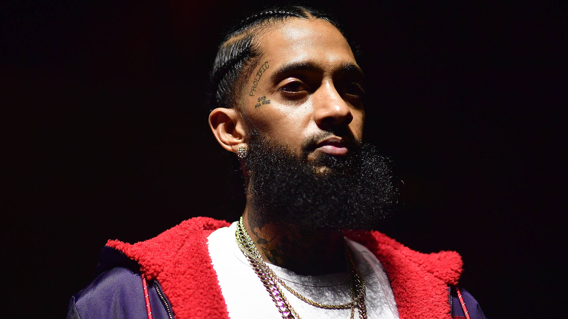 傳奇饒舌藝術家Nipsey Hussle登台演出 壁紙 — 在戲劇性舞台燈光下，一名留著濃密鬍鬚和紋身的男子的特寫肖像。
