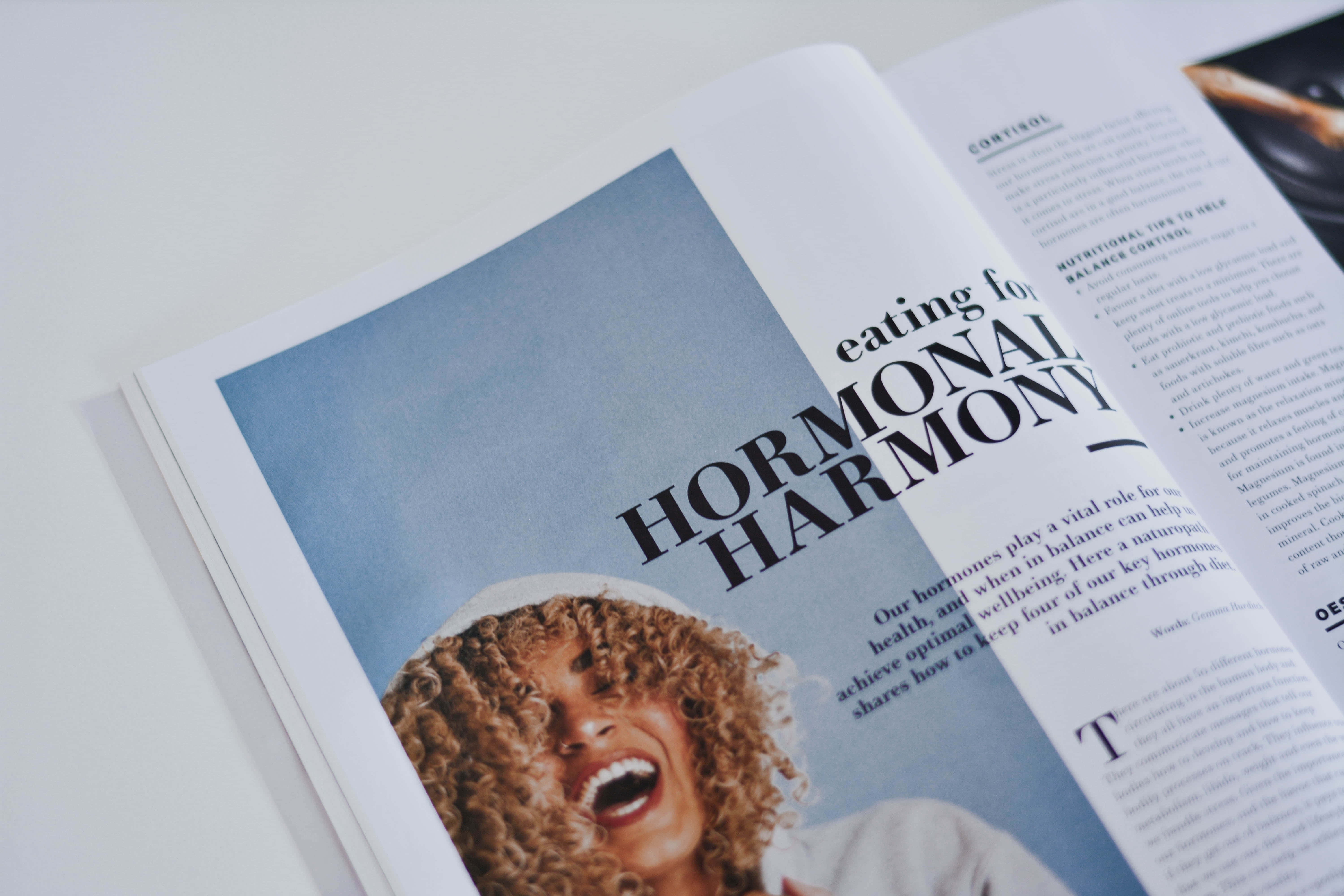 傳播新聞！ 壁紙 — 一位快樂的捲髮女性在名為「Hormonal Harmony」的雜誌跨頁上，對著柔和的藍色背景開懷大笑。