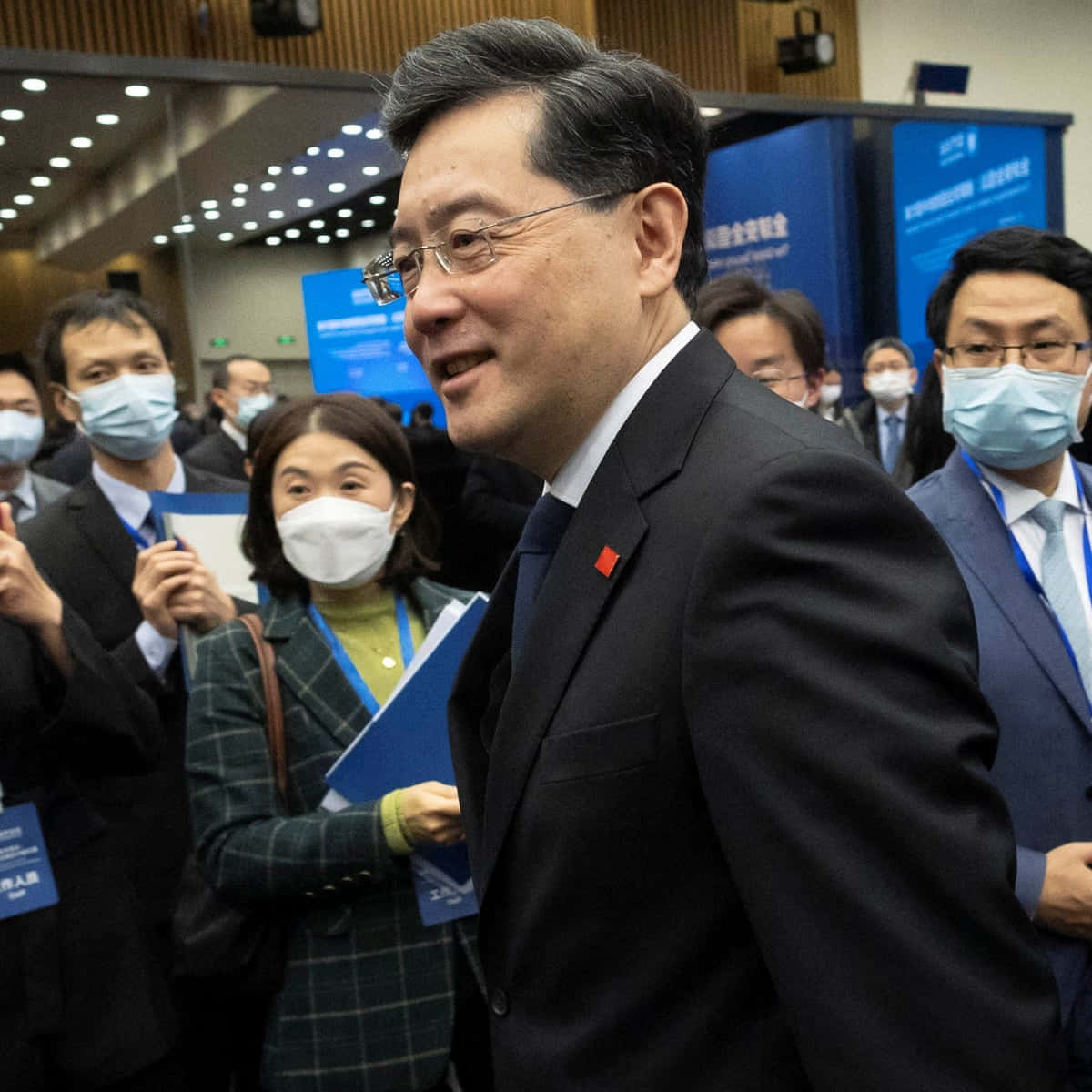 傳統中式橋樑與寧靜河景 壁紙 — 一位微笑的男子身穿深色西裝和眼鏡，在室內穿過一群穿著正式並佩戴口罩的人群。