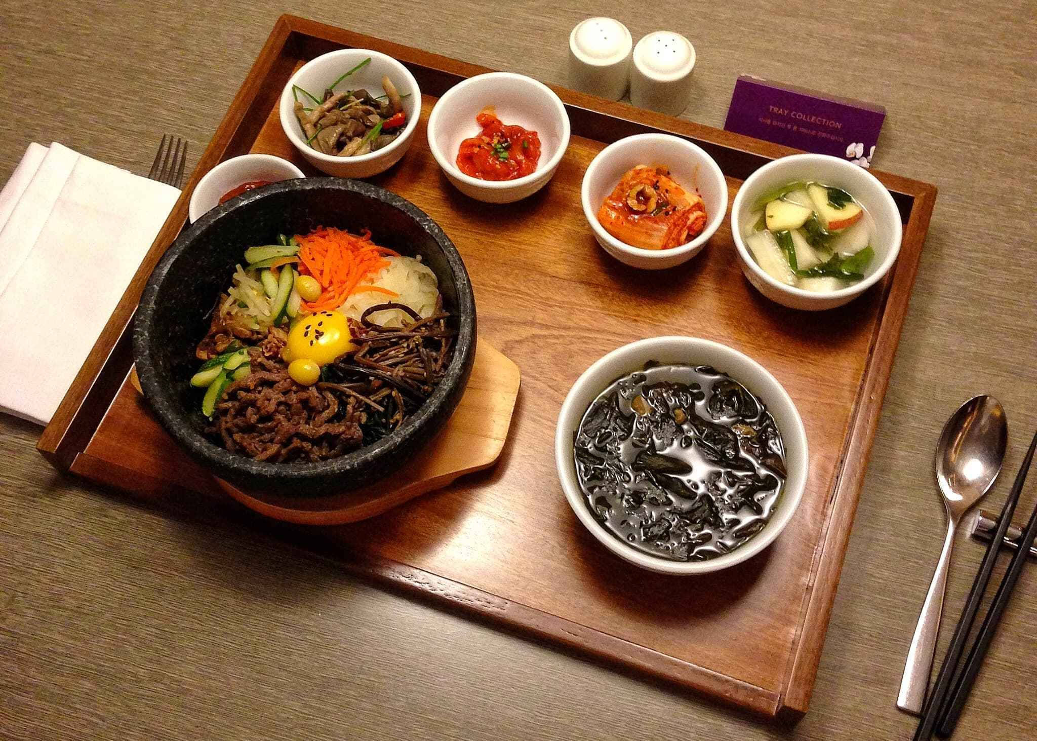 傳統全州拌飯 壁紙 — 木托盤上放著一個滋滋作響的石鍋拌飯，配有色彩繽紛的蔬菜和牛肉，周圍有配菜和湯。