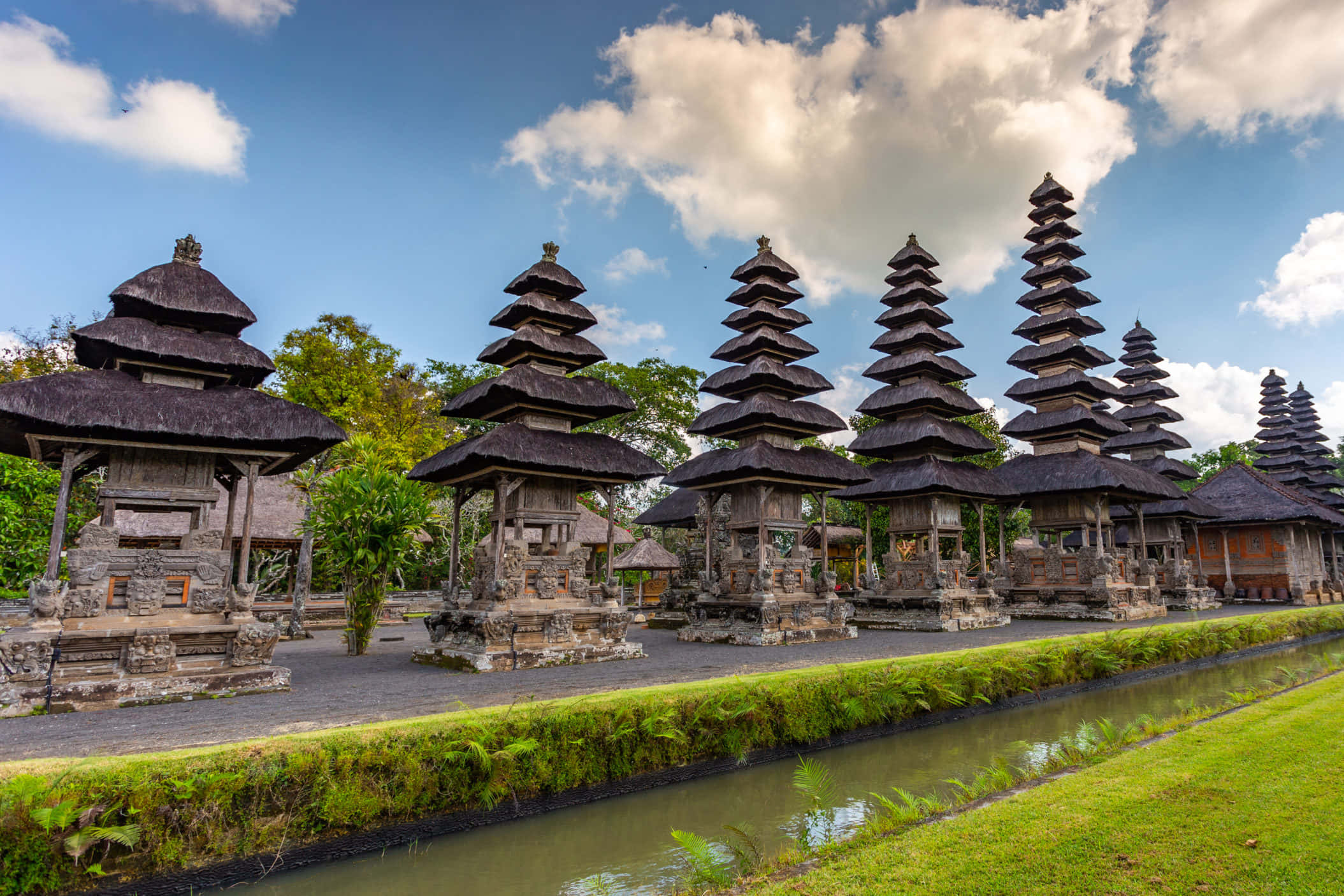Indonesia 桌布 壁紙 — 傳統巴厘島寺廟建築，階梯式茅草屋頂排列在寧靜的水道旁，上方是晴朗的藍天和白雲。