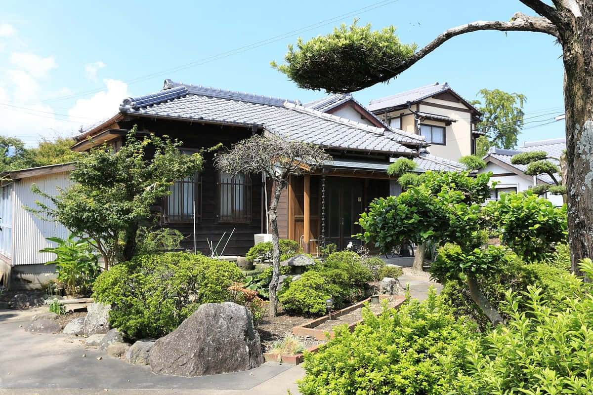 Samurai Garden 桌布 壁紙 — 傳統日式木屋，瓦片屋頂，坐落於茂密綠色花園中，上方是明亮的藍天。