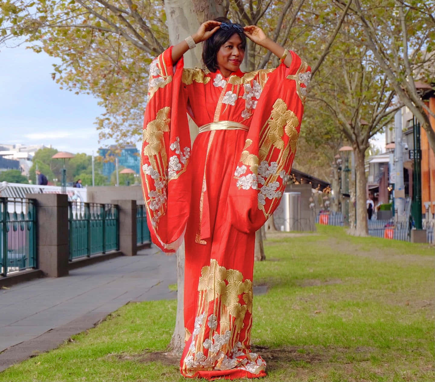 傳統日本和服 壁紙 — 一位身穿鮮紅色和服、飾有金色和白色花卉刺繡的女性，在陽光明媚的城市公園中，優雅地站在枝葉繁茂的樹下。