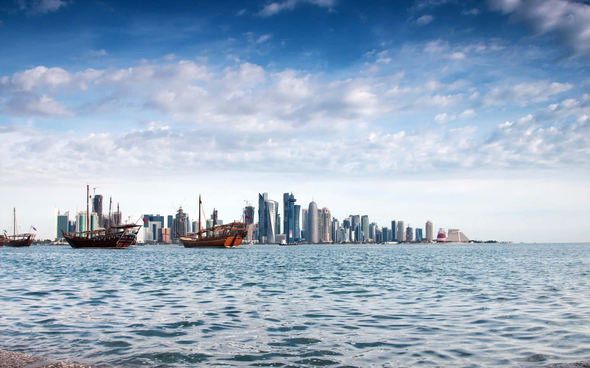 Qatar 桌布 壁紙 — 傳統木製單桅帆船在平靜的藍色水面上漂浮，背景是部分多雲的天空下現代摩天大樓的天際線。