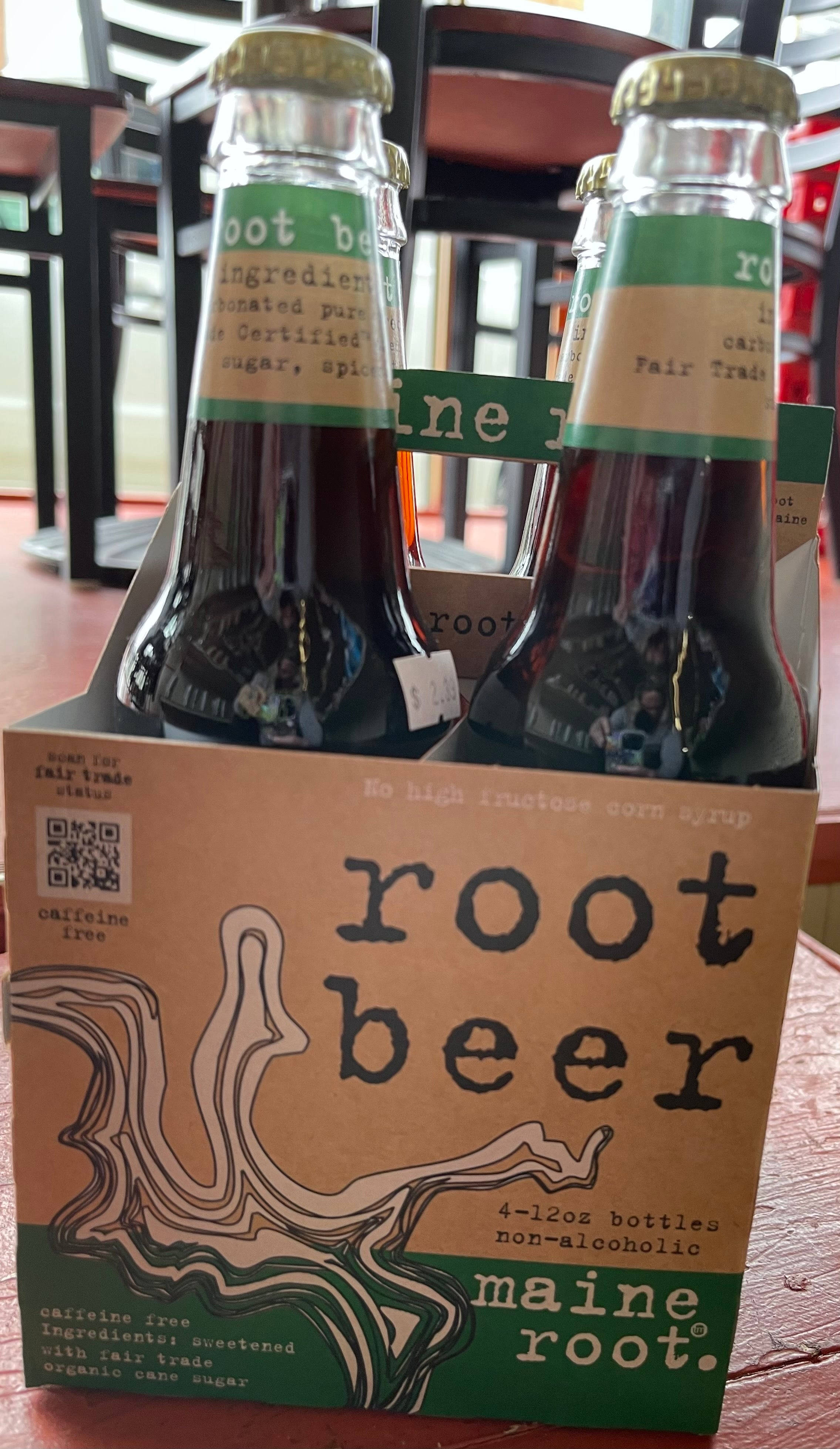 傳統緬因根根啤酒禮盒 壁紙 — 一個裝有Maine Root根汁啤酒瓶的紙板六連包放在木桌上，背景為模糊的室內環境。