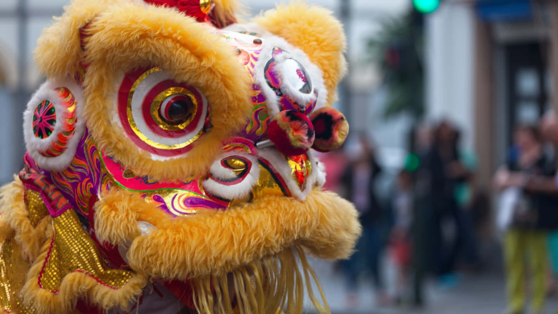 傳統舞獅服裝 壁紙 — 鮮黃色舞獅服裝，帶有複雜的紅色、金色和粉紅色圖案，在模糊的城市街景中脫穎而出。