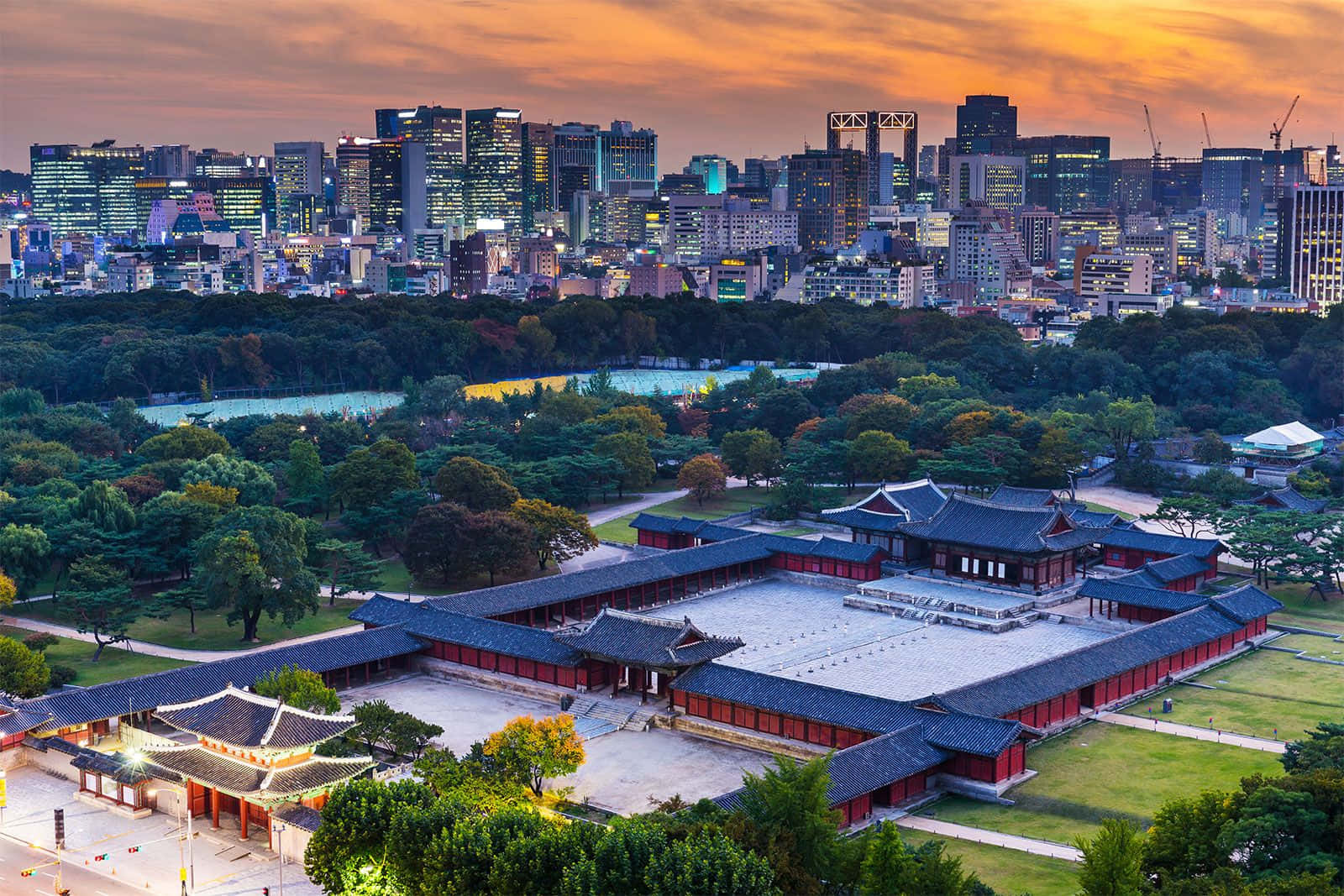 South Korea 桌布 壁紙 — 傳統韓國宮殿，紅牆黑瓦，坐落在綠色公園中，以夕陽下閃耀的現代城市天際線為背景。
