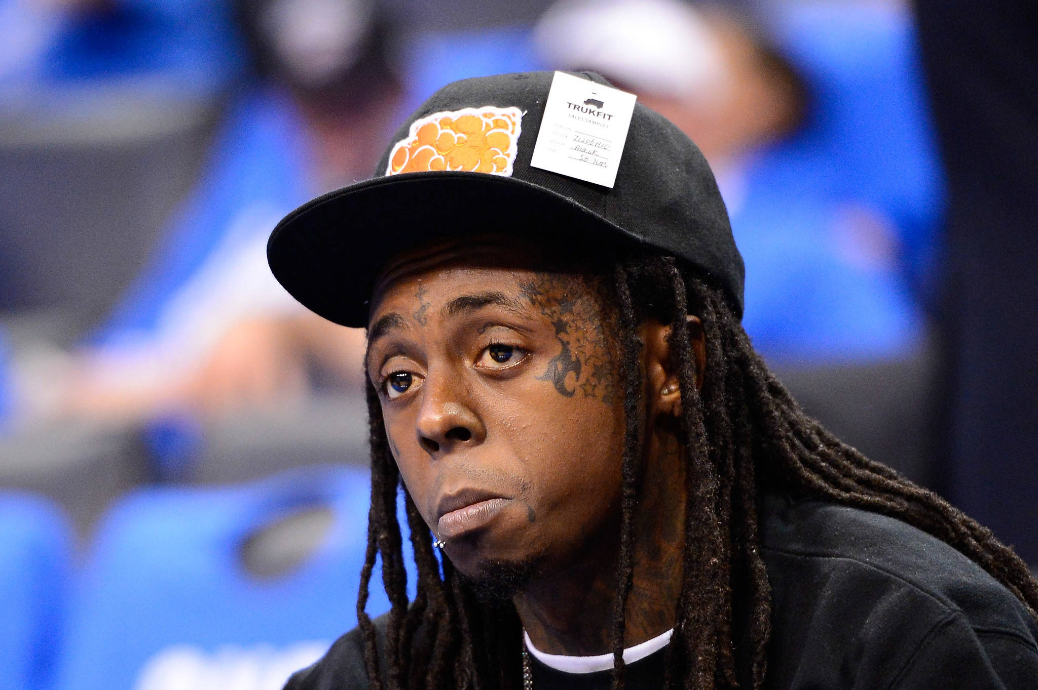 傷感的 Lil Wayne 壁紙 — 饒舌歌手Lil Wayne的close-up肖像，戴著黑色帽子，上有橙色標誌和白色標籤，背景是模糊的藍色體育場座位。