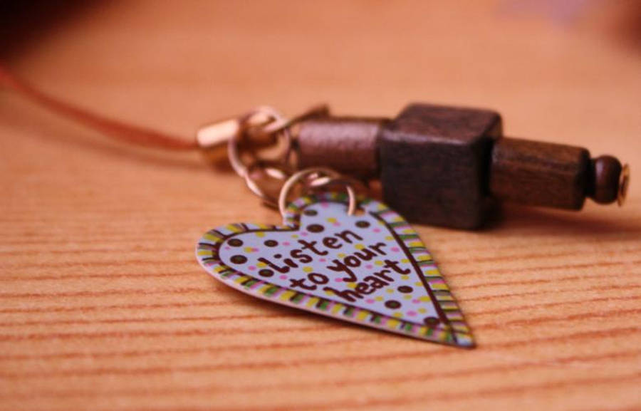 傾聽你的心格言 壁紙 — 一個手工製作的愛心吊飾，上面寫著「listen to your heart」，掛在一條木珠項鍊上，放在溫暖的木質表面上，特寫鏡頭。