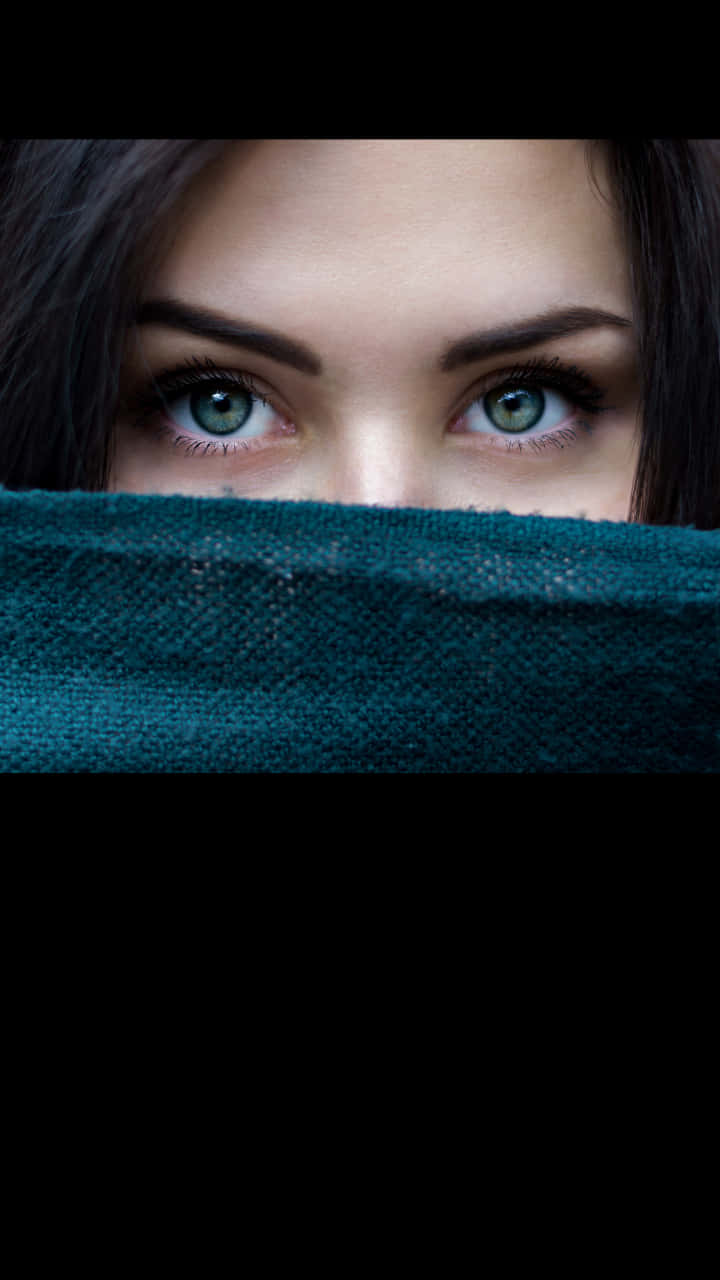 女人綠色眼睛從藍綠色圍巾上方窺視，背景為黑色。