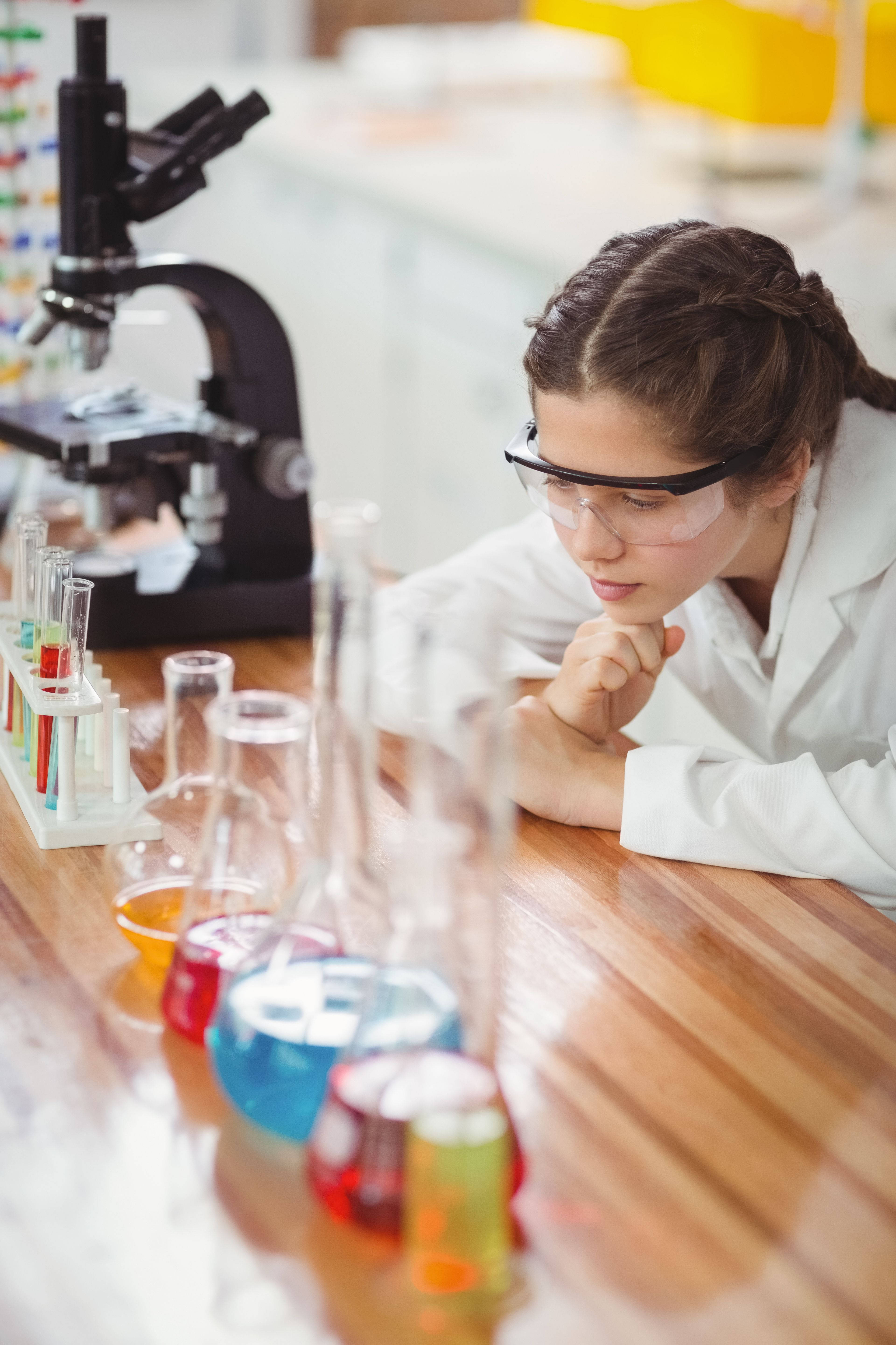 像科學家一樣學習 壁紙 — 一位專注的年輕科學家穿著實驗室外套和安全護目鏡，俯身在一張木桌上，桌上擺滿了裝有彩色液體的燒瓶和試管。
