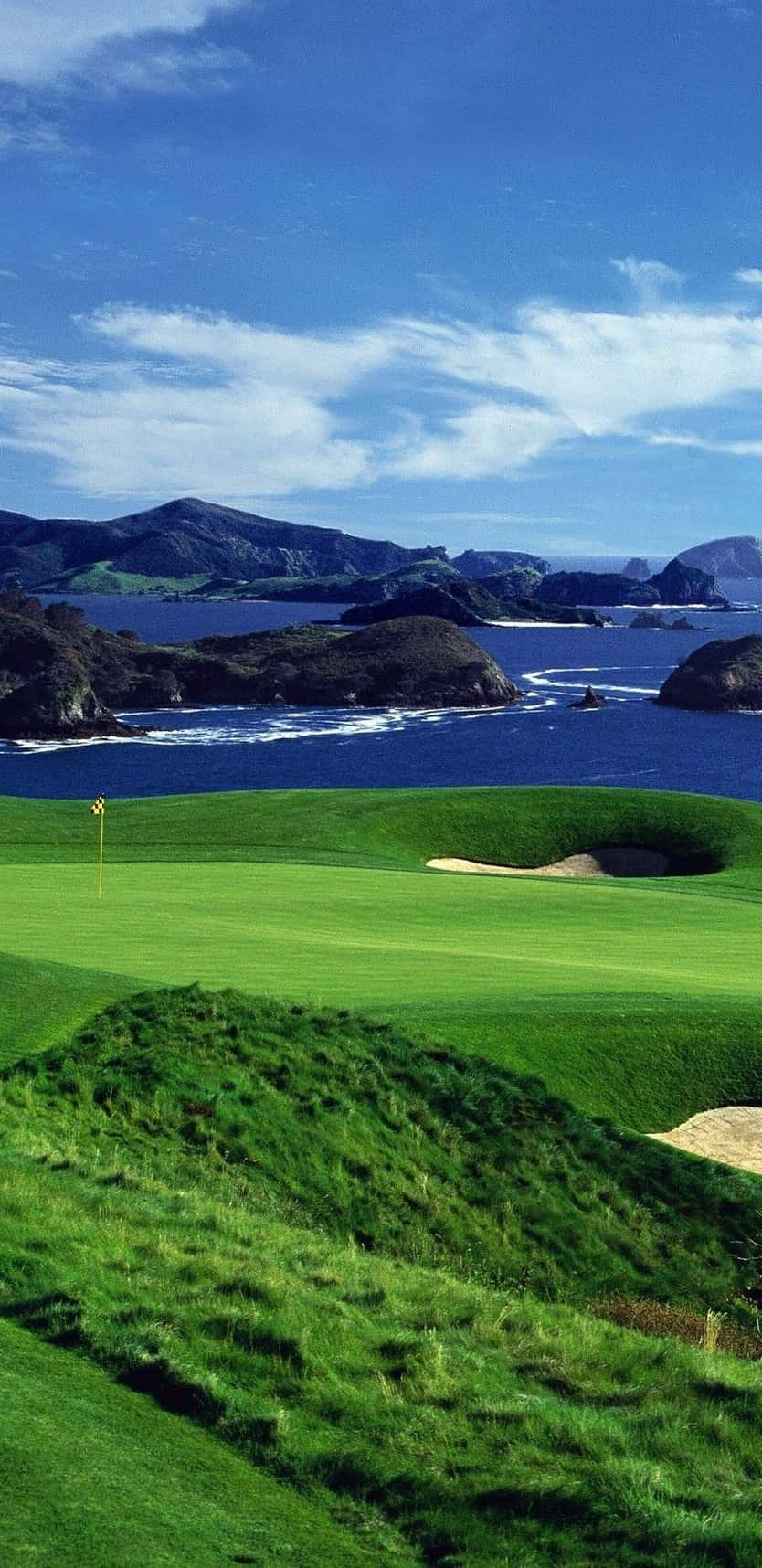 像素3考瑞崖高爾夫球場背景 壁紙 — 一個充滿活力的綠色高爾夫球場擁抱著崎嶇的海岸線，深藍色的海洋和遠處的山脈在明亮的藍天下。