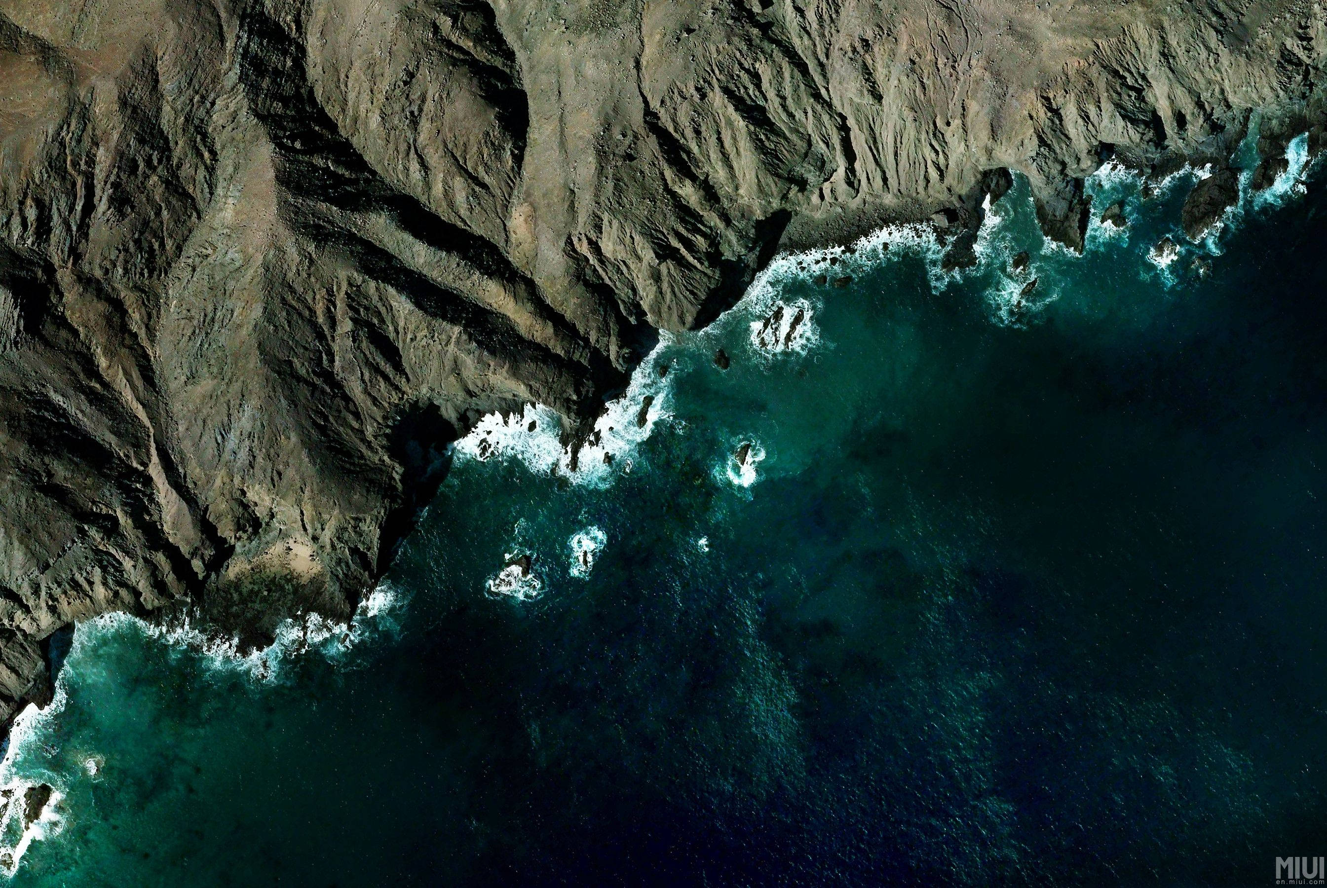 像素海灘與山脈風景 壁紙 — 崎嶇懸崖與碧綠海洋交匯，白色波浪沿著鋸齒狀海岸線拍打的鳥瞰圖。