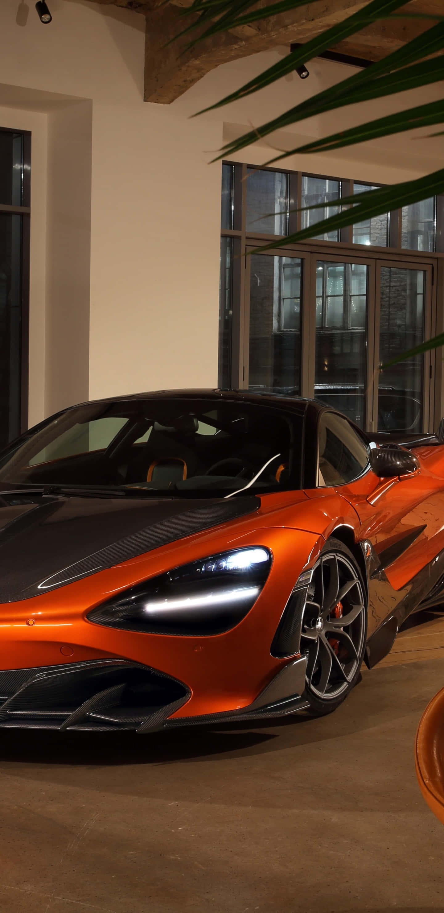 像素風橙色麥拉倫720S壁紙 壁紙 — 一架時尚的橙色McLaren跑車在溫暖的室內燈光下，在大窗戶和綠色棕櫚葉旁閃閃發光。