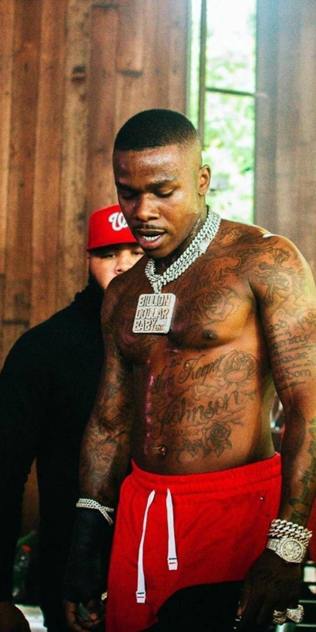 億萬寶貝 DaBaby 手機 壁紙 — 一名紋身男子穿著紅色短褲和鑽石鏈，赤膊站在室內，身後有木牆和窗戶。