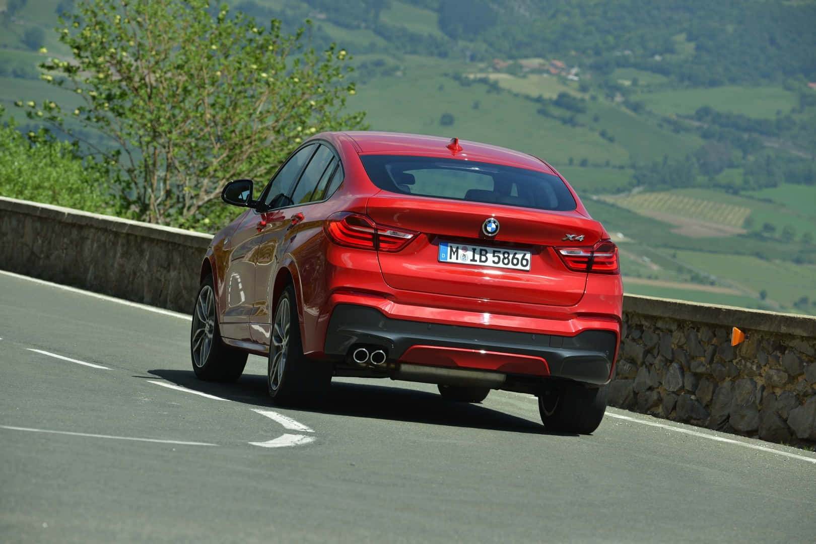 優雅姿態下的 BMW X4：時尚與性能的完美結合 壁紙 — 一輛亮面紅色BMW X4在明媚日光下行駛於蜿蜒的山路上，兩旁是翠綠的山丘和石牆。