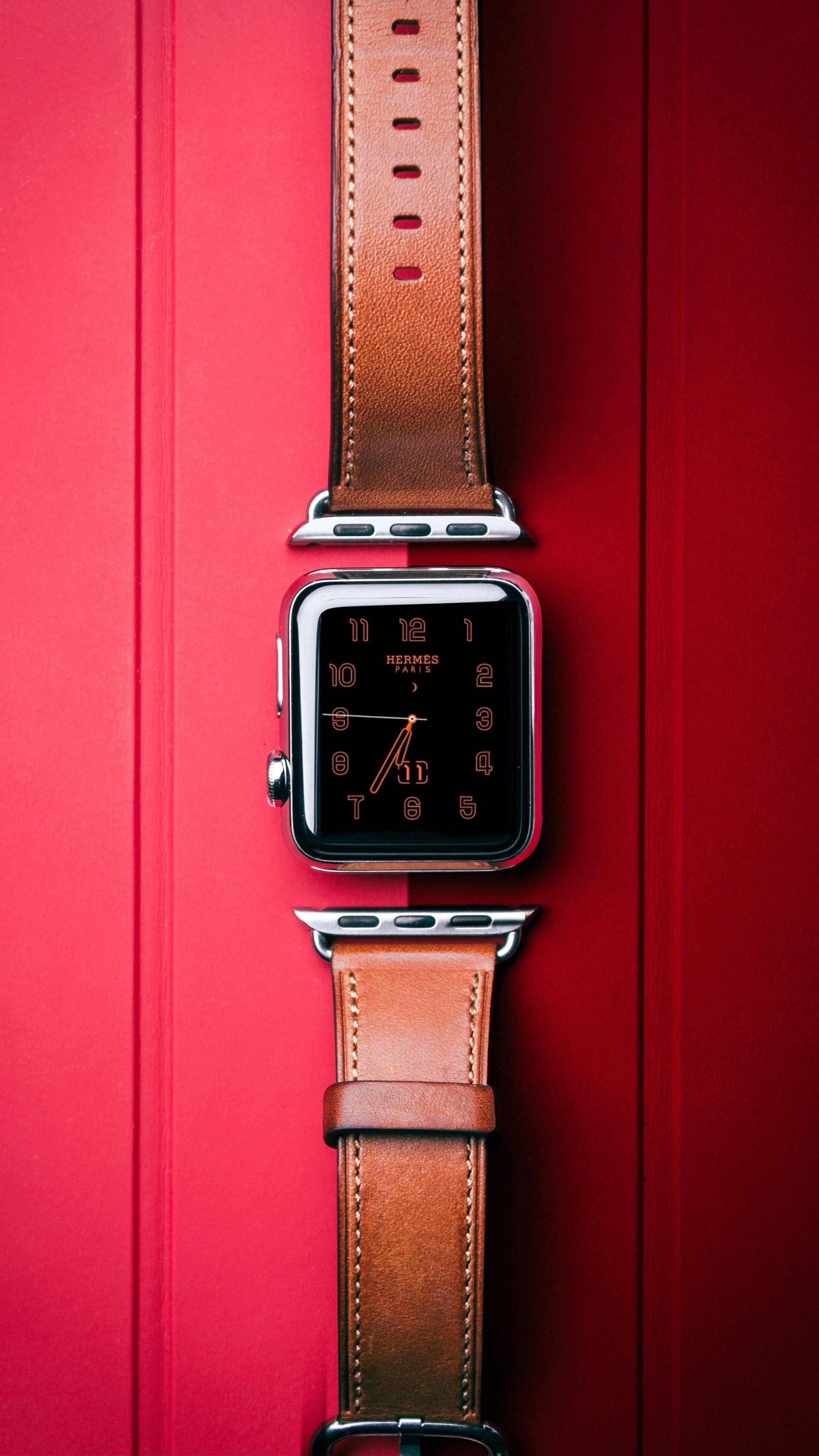 優雅愛馬仕手錶 壁紙 — 一款時尚的Apple Watch搭配棕色皮革Hermès錶帶，垂直放置在鮮明的紅色紋理背景上。