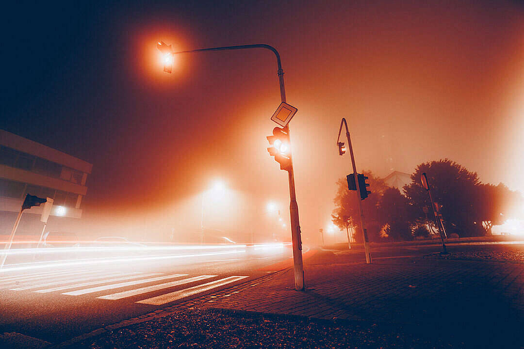 優雅淺棕交叉路口交通燈 壁紙 — 霧氣瀰漫的夜晚街道，被發光的橙色街燈照亮，汽車燈光劃過斑馬線形成光軌。