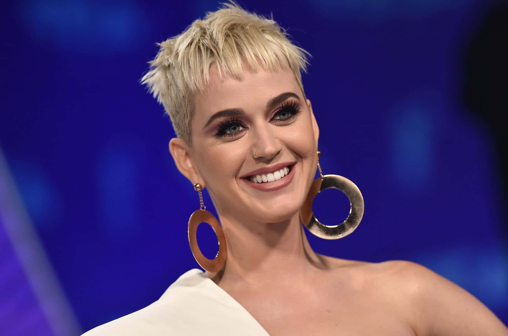 優雅珍珠耳環 壁紙 — Katy Perry 在藍色背景前，留著短金髮、戴著大圓耳環，露出燦爛笑容。