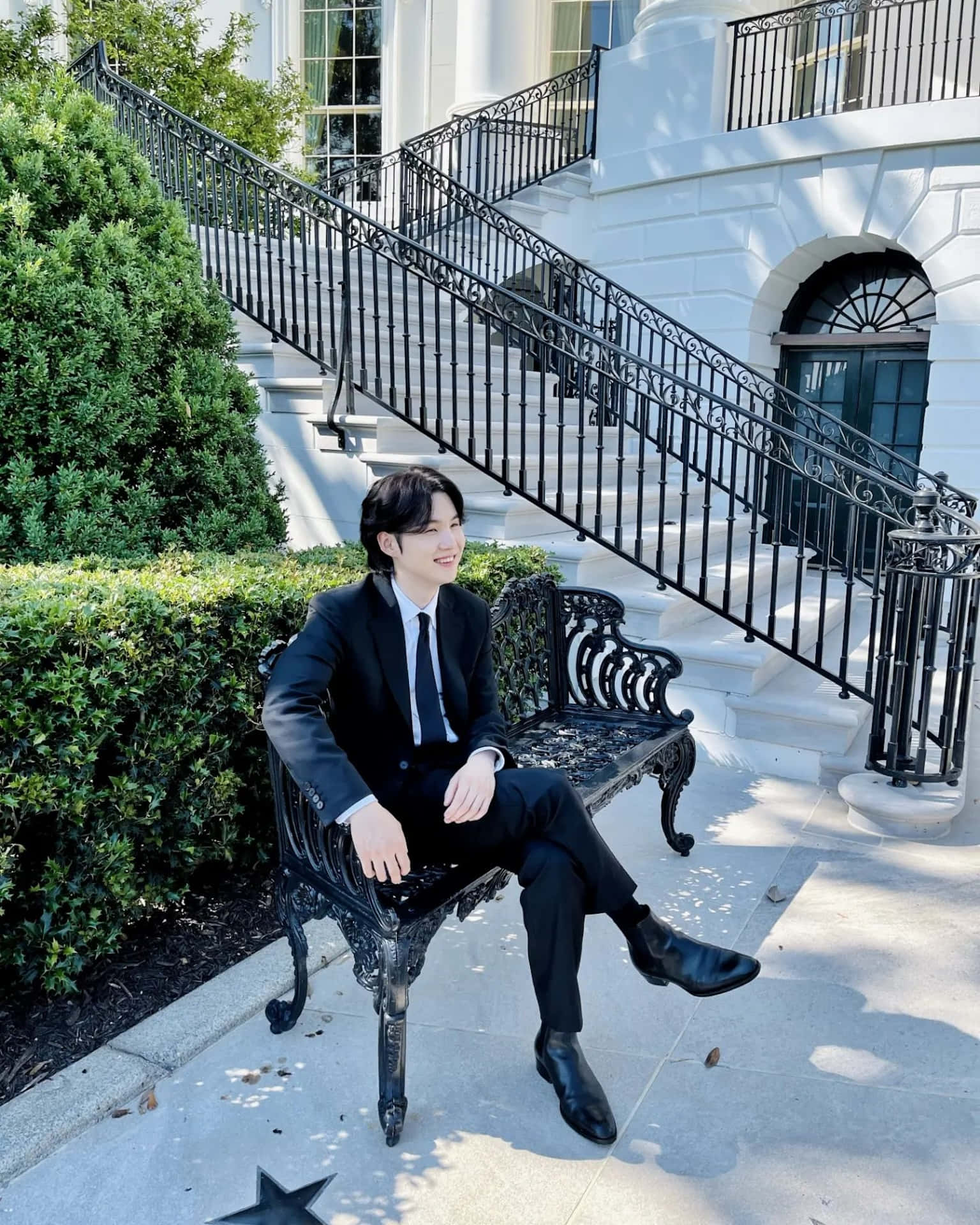優雅男士坐於戶外階梯旁 壁紙 — 一名身穿俐落黑色西裝的男子，放鬆地坐在一座宏偉白色大宅前裝飾華麗的長椅上，大宅有寬闊的樓梯和茂密的綠色樹籬。