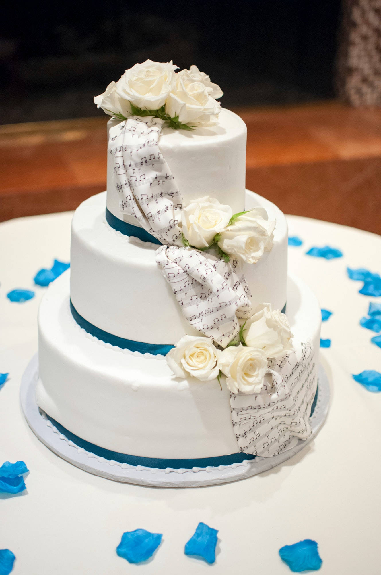 優雅白玫瑰多層婚禮蛋糕 壁紙 — 三層白色結婚蛋糕，飾有白玫瑰和樂譜絲帶，周圍散落著藍色花瓣在白色桌子上。