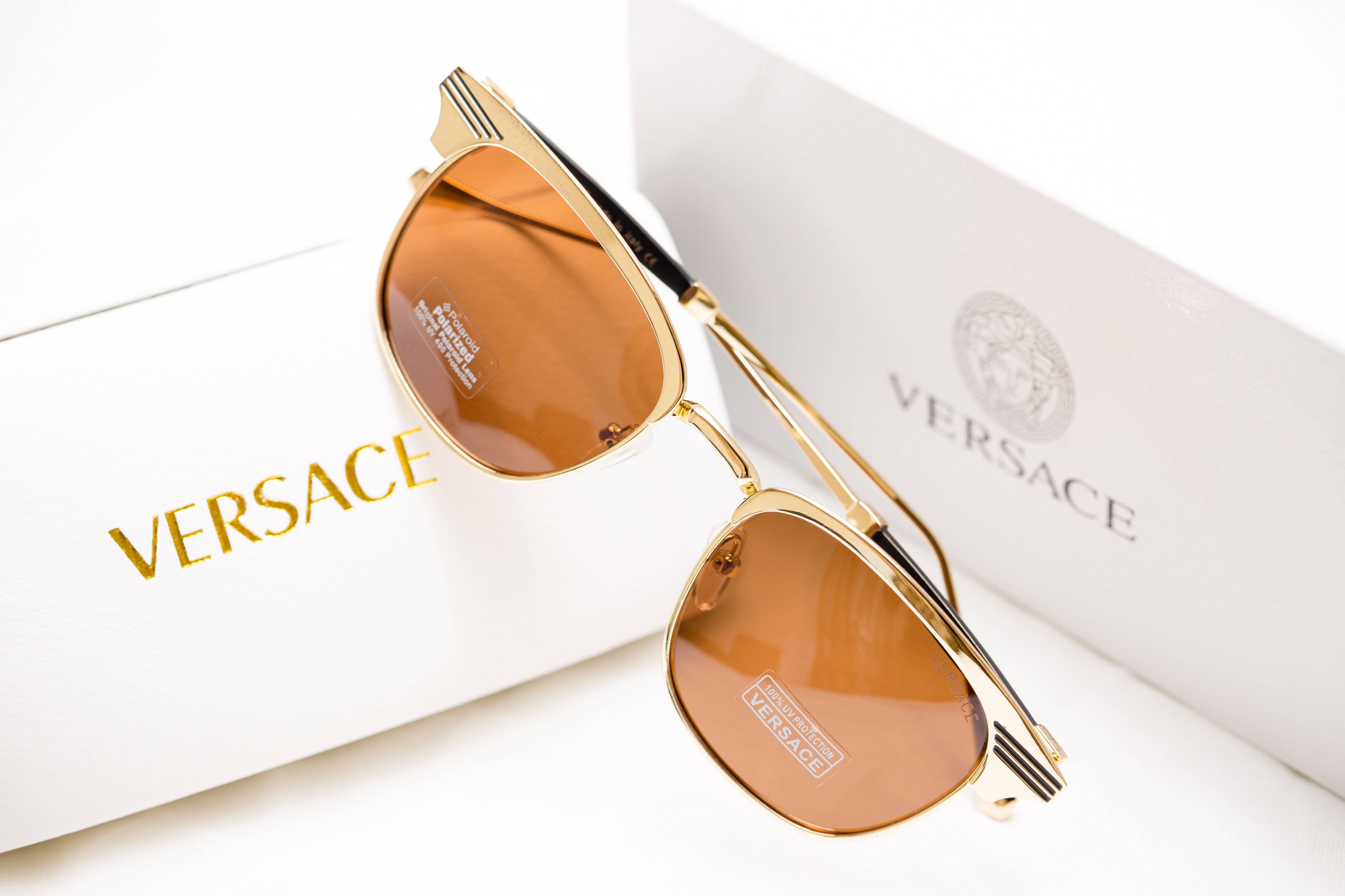 優雅的 Versace 棕色太陽眼鏡，為您的服裝增添高雅氣質 壁紙 — 金色框Versace太陽眼鏡配琥珀色鏡片，優雅地放置在品牌白色包裝上，背景為純白色。