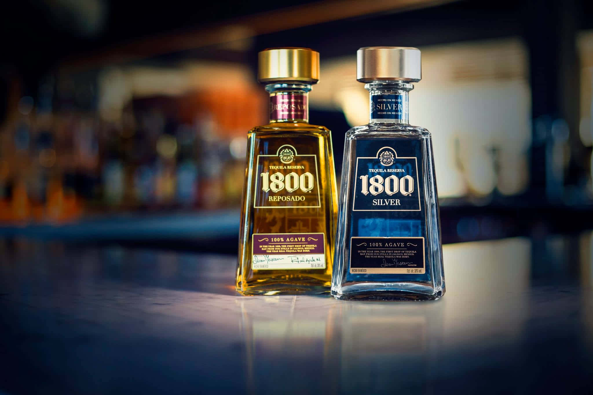 優雅的1800龍舌蘭酒銀色與陳年版本 壁紙 — 兩瓶琥珀色和銀色的1800龍舌蘭酒放在拋光吧檯上，背後是模糊的酒架。