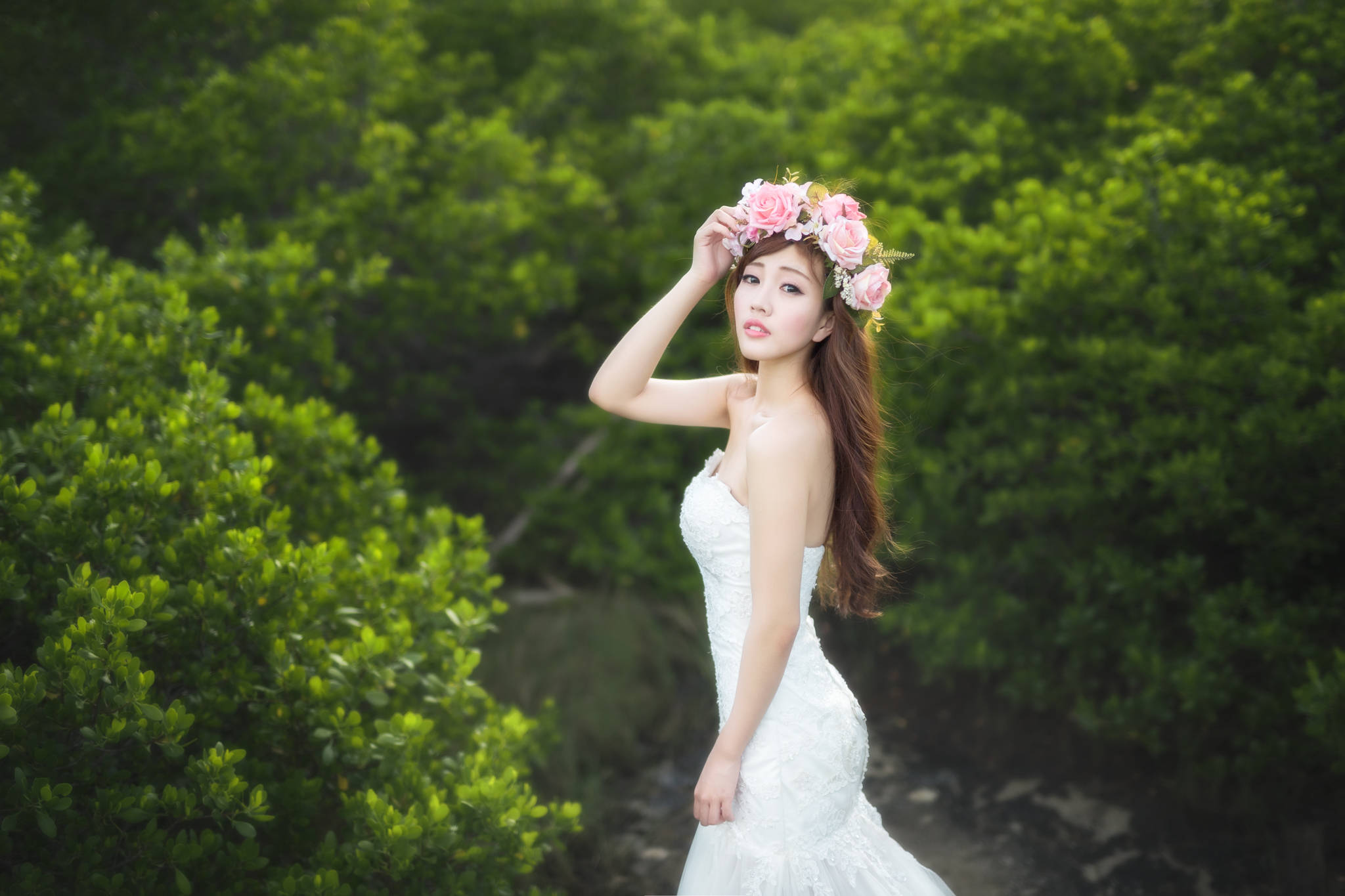 優雅的化身：燦爛新娘 壁紙 — 一位身穿白色連衣裙的女性戴著粉紅色花冠，在茂密的綠色灌木叢中擺姿勢。