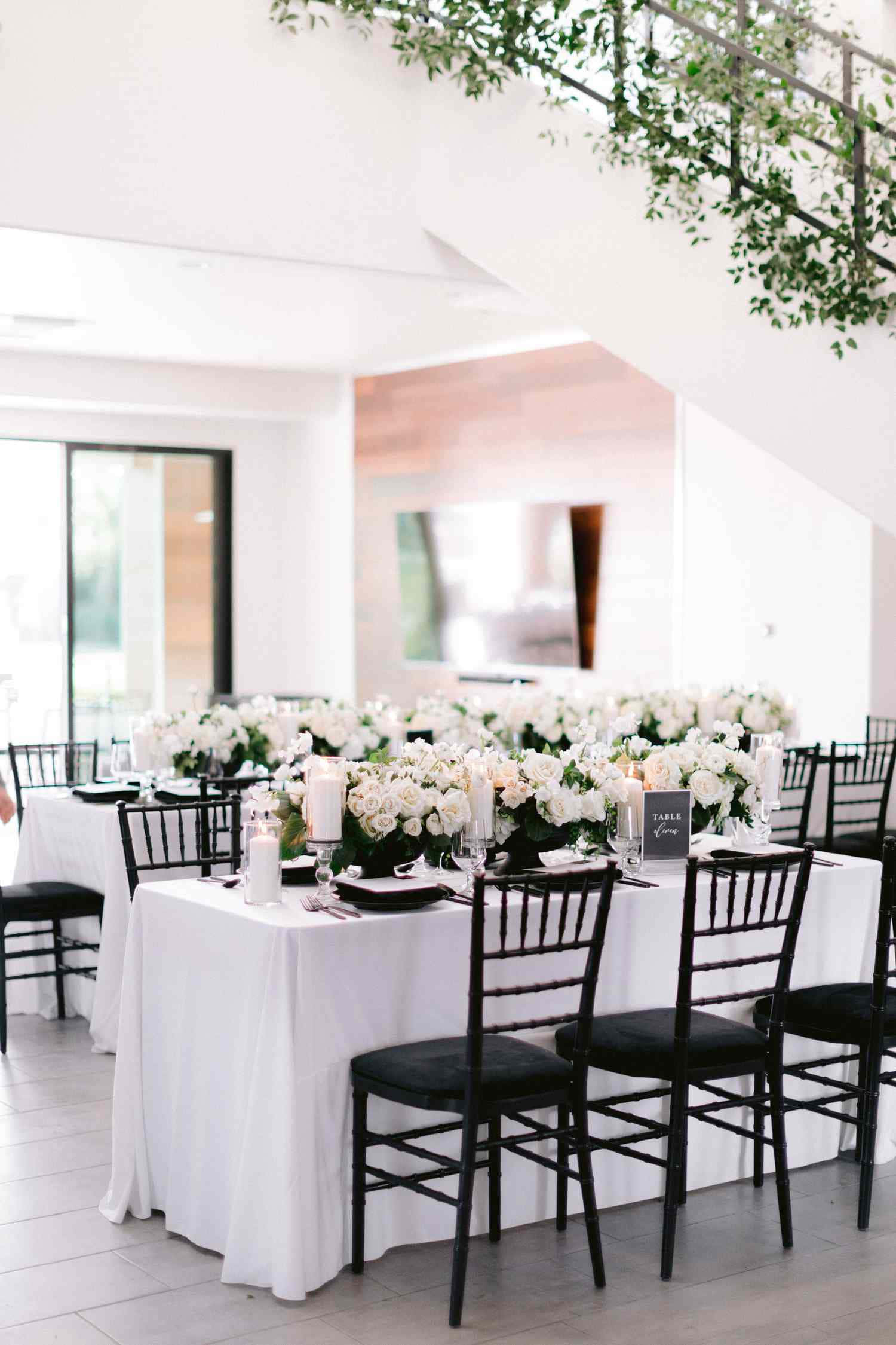 Black And White Wedding 桌布 壁紙 — 優雅的婚宴餐桌鋪上白色亞麻布，配黑色椅子，在懸掛的綠色植物下擺放茂盛的花卉中心裝飾。