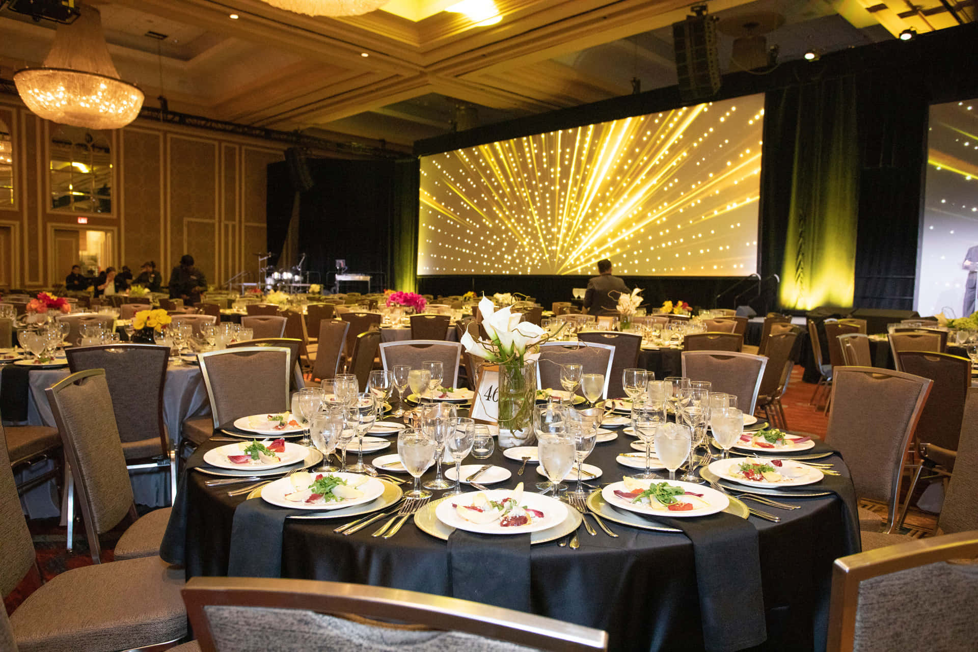 Corporate Event 桌布 壁紙 — 優雅的宴會廳，圓桌已為正式活動佈置好，在溫暖的水晶吊燈和耀眼的金色星爆屏幕下。