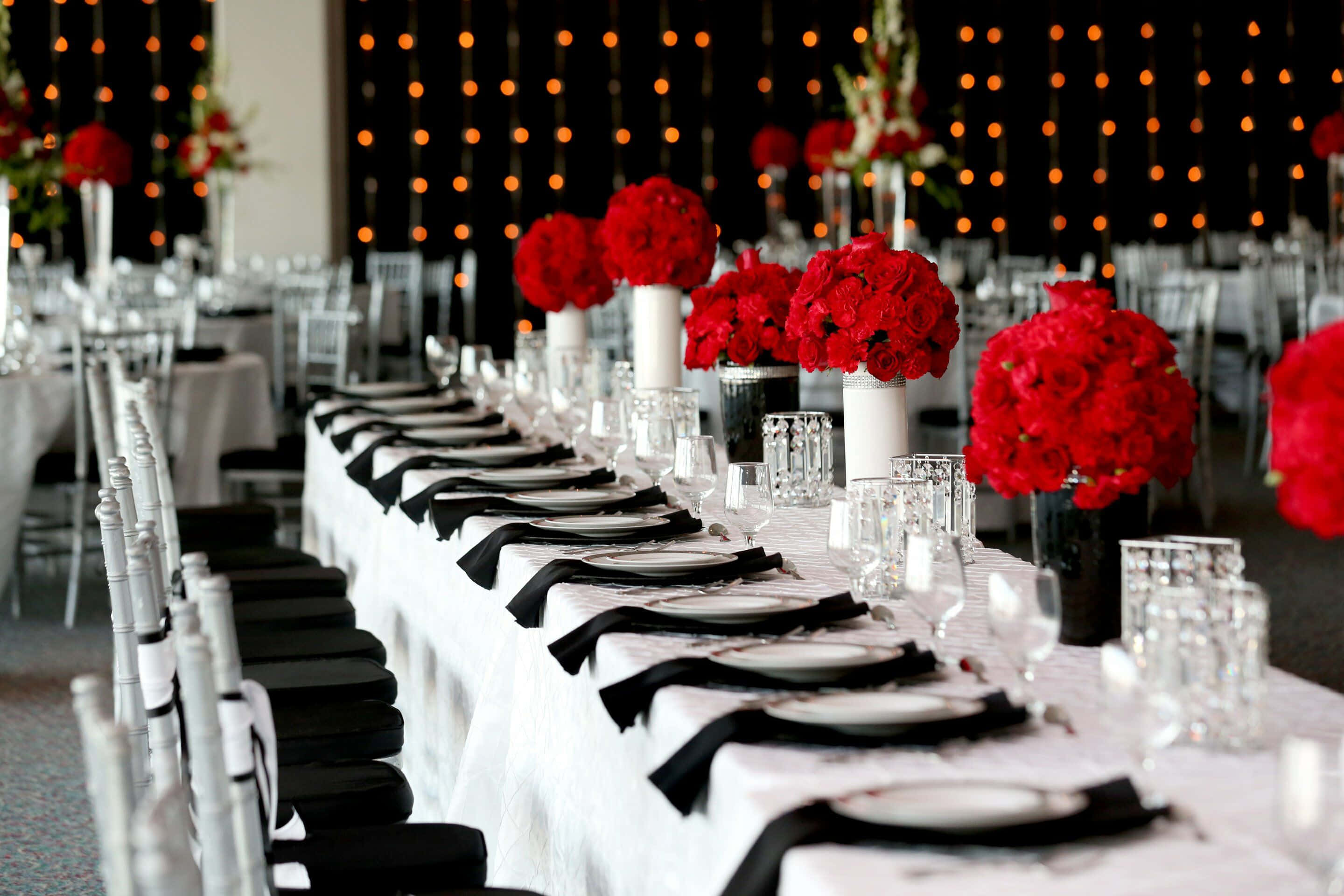 Black And White Wedding 桌布 壁紙 — 優雅的宴會餐桌，鋪有白色亞麻布、黑色餐巾和鮮豔的紅色花卉中心裝飾，在溫暖的串燈下。