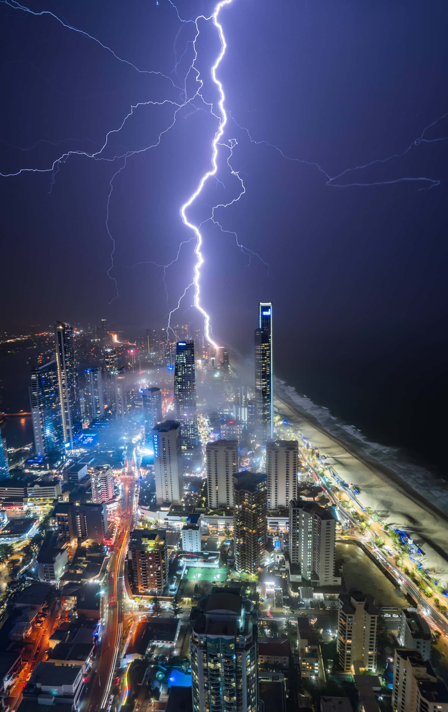 優雅的暴風雨夕陽景色 壁紙 — 一道巨大的閃電劃過充滿活力的夜間海岸城市景觀，閃耀的摩天大樓和燈火通明的海灘。