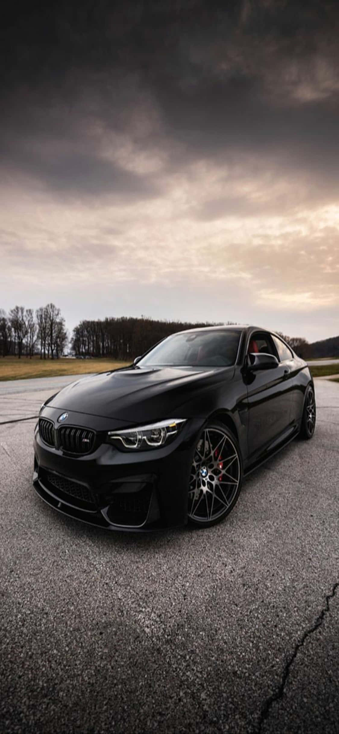 優雅的結合：iPhone X 與 BMW 壁紙 — 一架時尚的黑色BMW M2雙門跑車停泊在裂開的瀝青路上，背景是黃昏時分戲劇性的多雲天空。