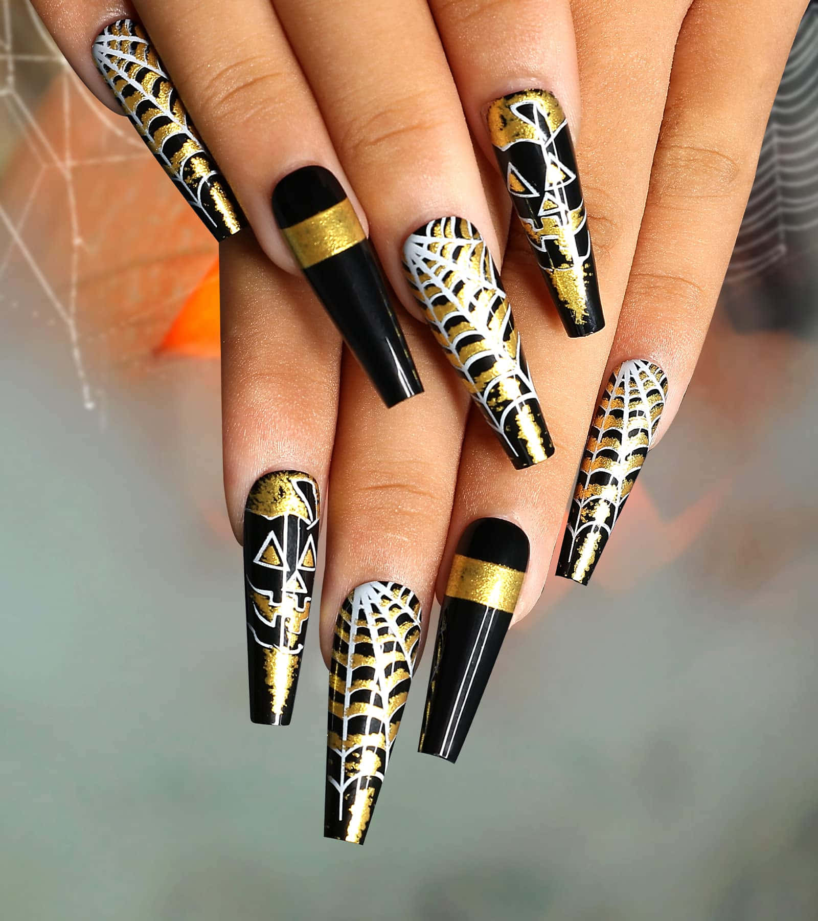 Halloween Nail Art 桌布 壁紙 — 優雅的黑金蜘蛛網圖案亞加力指甲，帶有幾何圖案，手部置於模糊背景前。